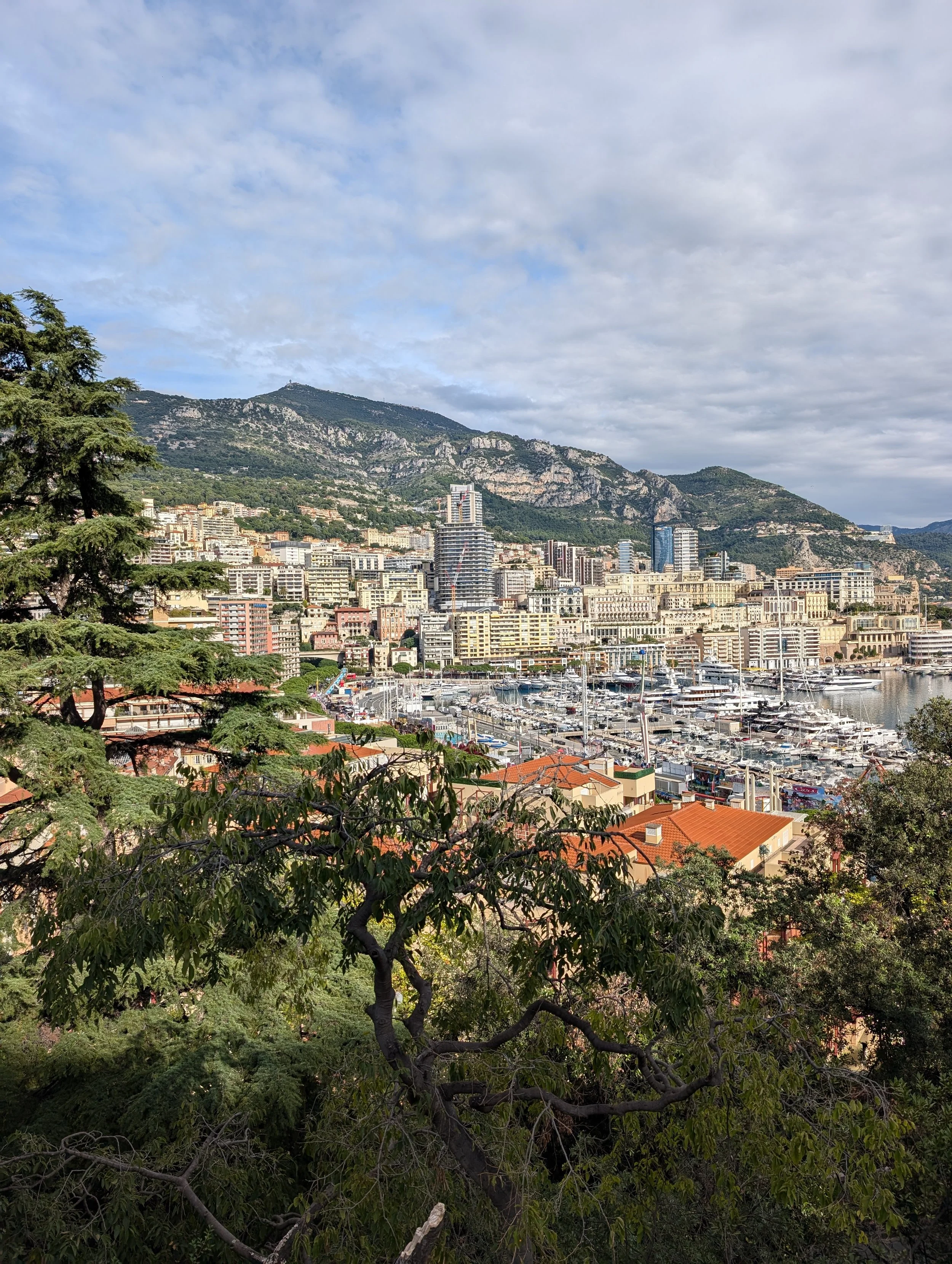 A city surrounded by mountain ranges with a marina of boats in the foreground and partly cloudy sky.