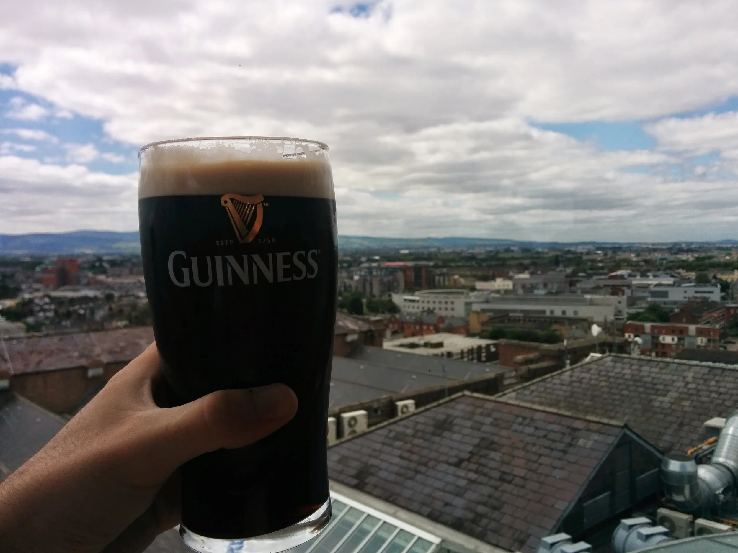 A hand holding a pint glass of Guinness stout beer with a cityscape and cloudy sky in the background.