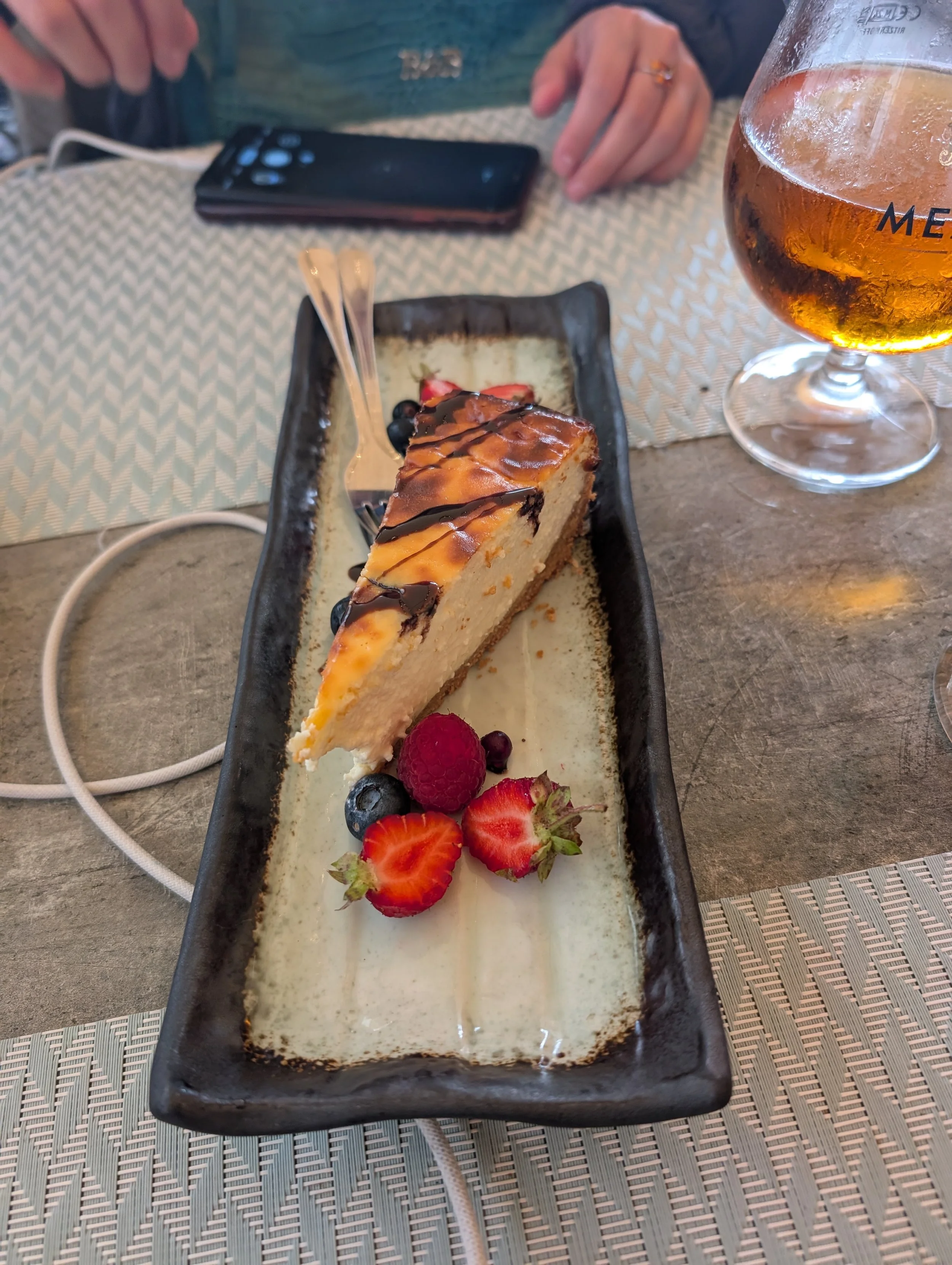 A slice of cheesecake topped with chocolate drizzle on a rectangular black plate, garnished with strawberries, raspberries, blackberries, and blueberries, with a glass of amber-colored beverage beside it on a table.