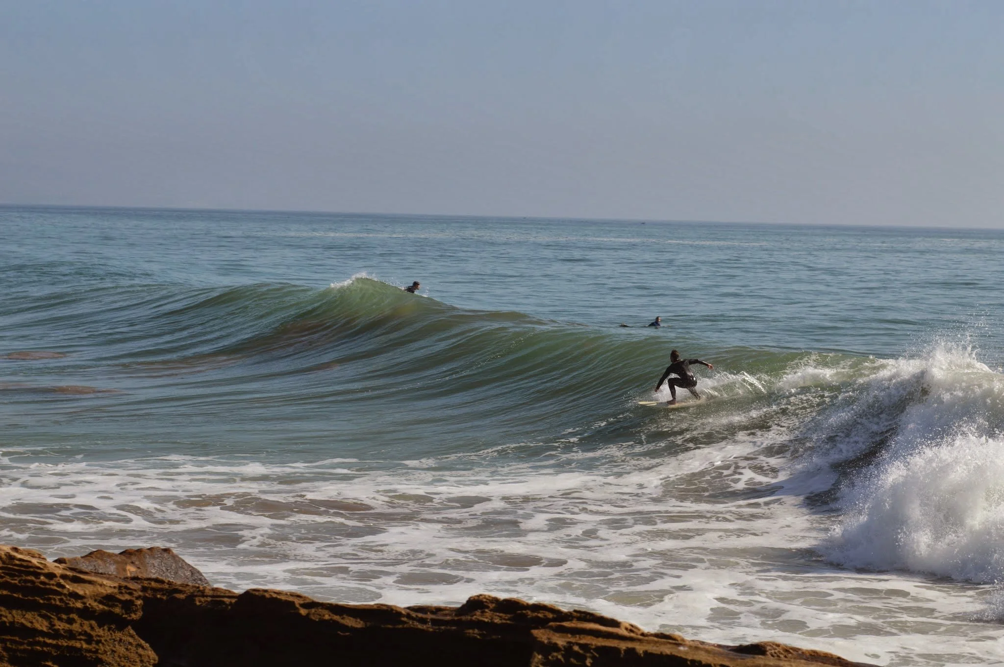 Surfers riding ocean waves near a rocky shoreline with a clear sky in the background.