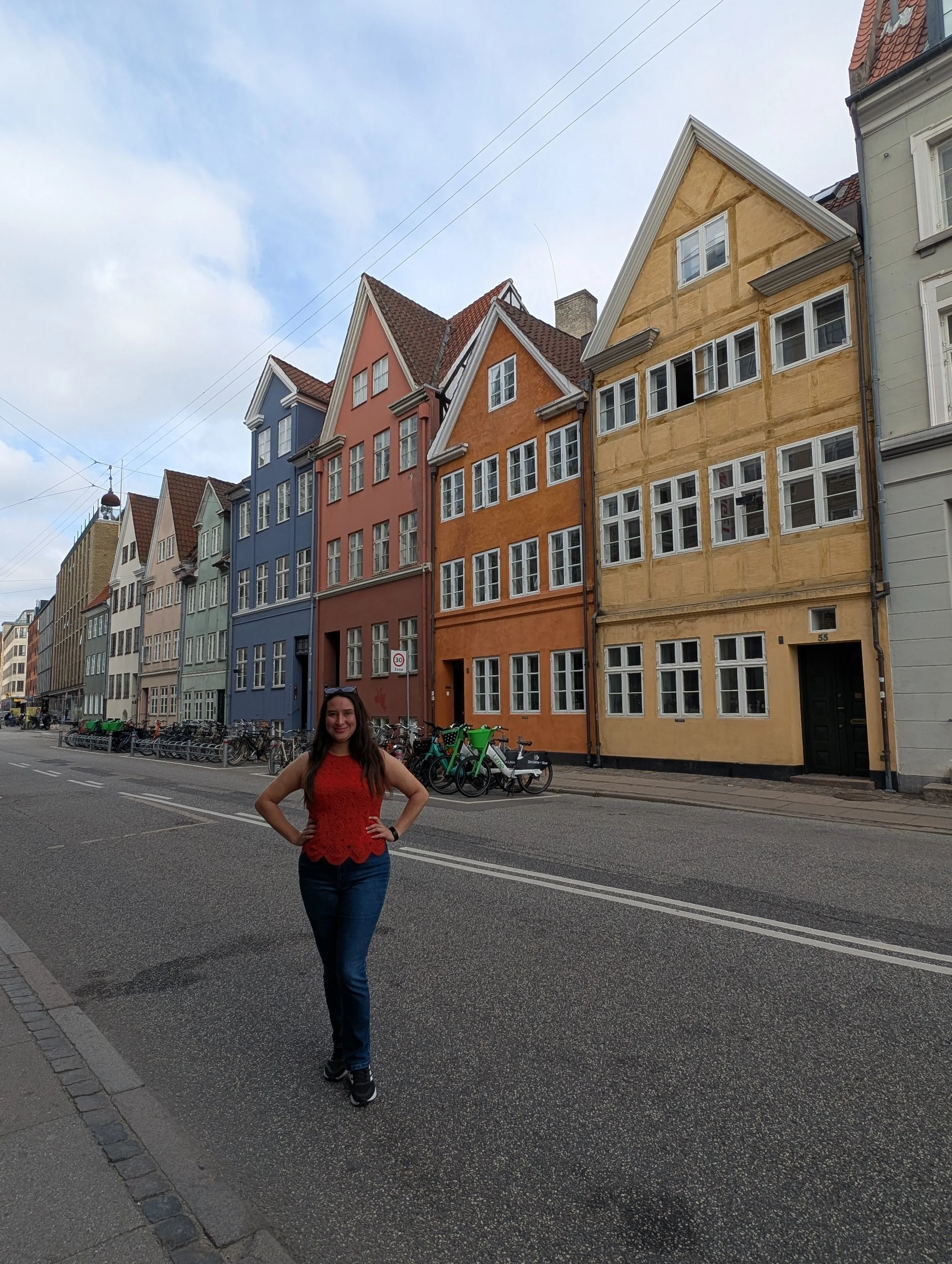 A young woman with long brown hair, wearing a red sleeveless top and blue jeans, standing on a city street with colorful buildings in the background.