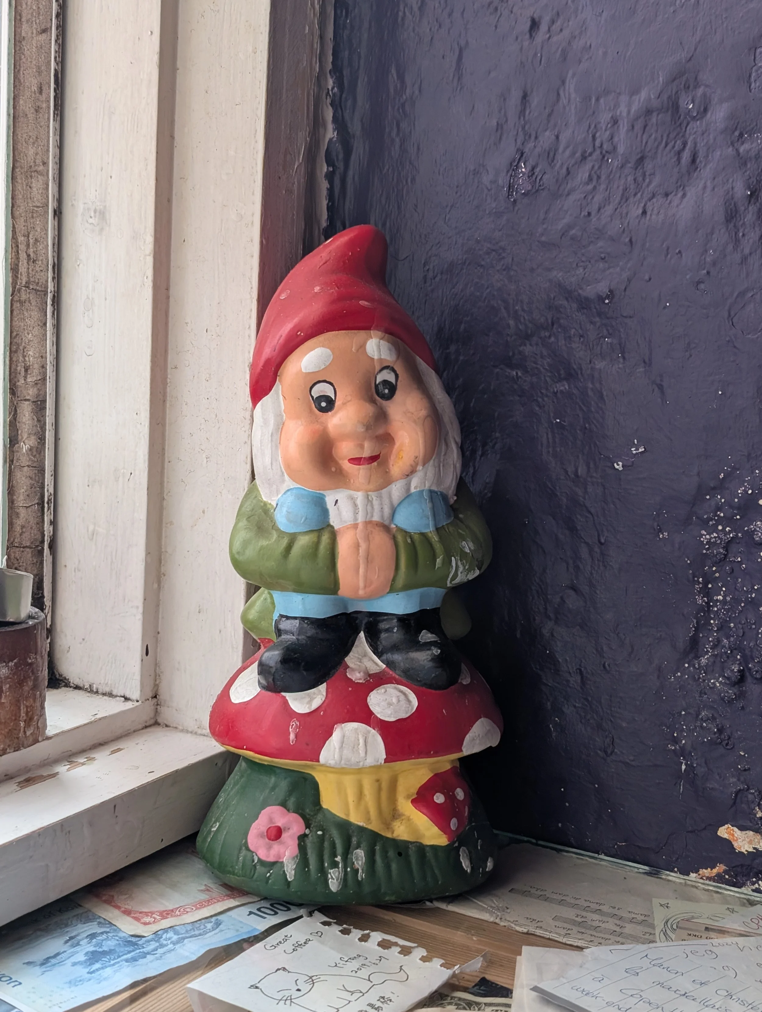 A colorful ceramic garden gnome with a red hat and blue shirt, standing on a mushroom-shaped base, placed on a windowsill next to some papers and drawings.