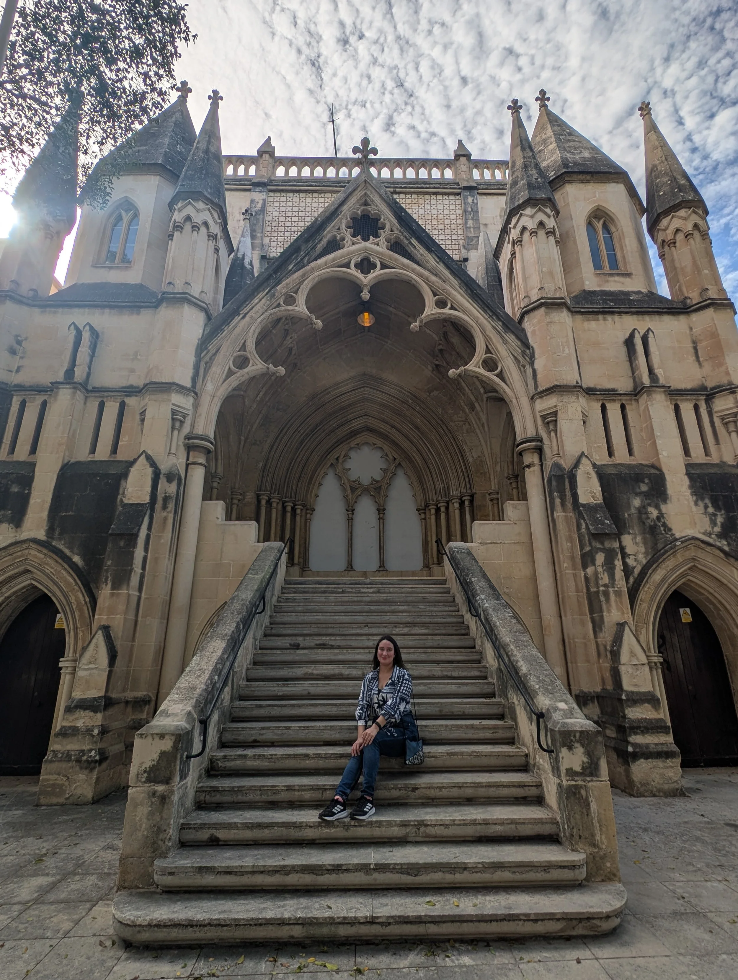 A woman sitting on the steps of a Gothic-style church with ornate architecture, tall spires, decorative arches, and stained glass windows, under a partly cloudy sky.