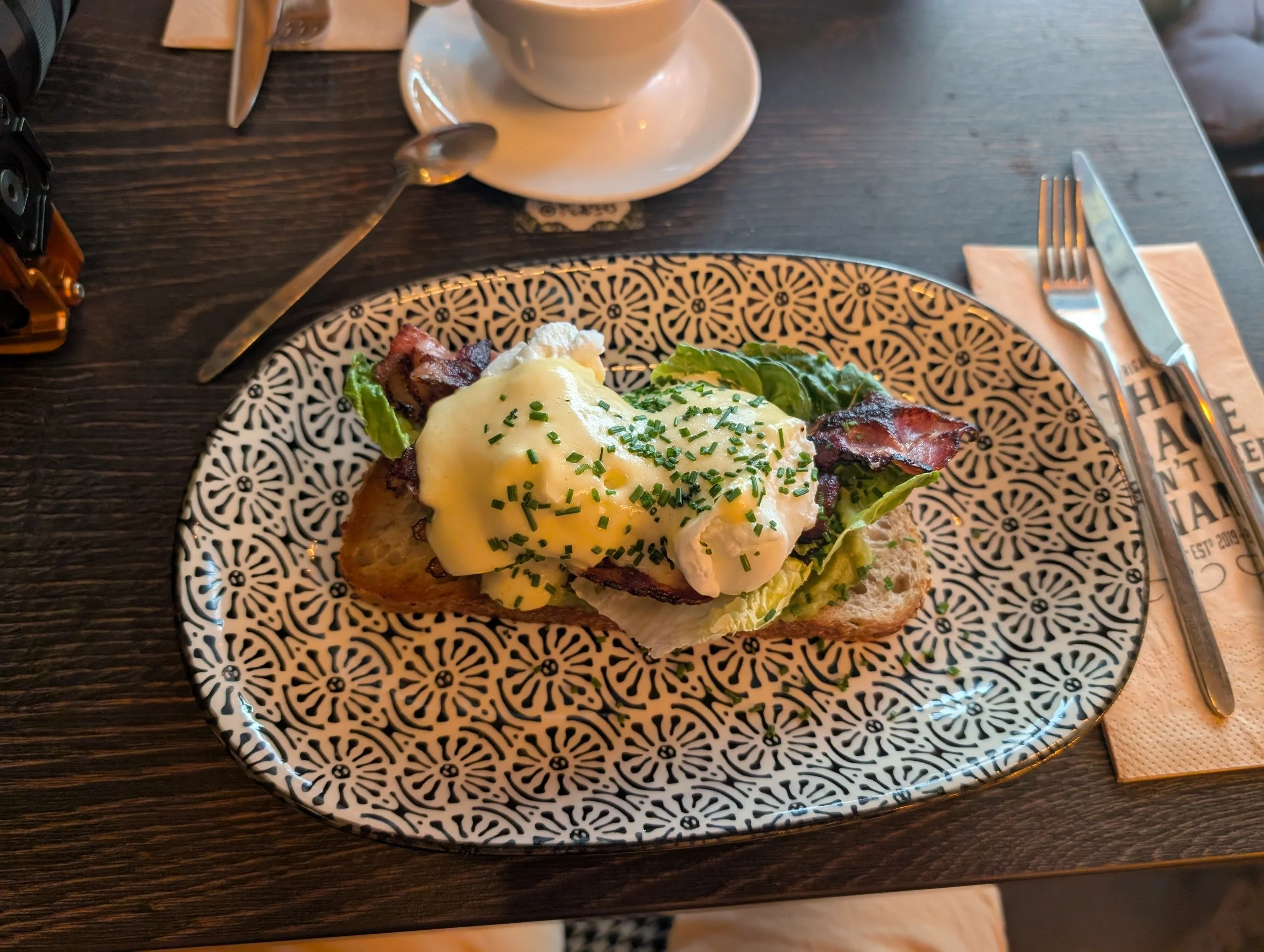A plate with an open-faced breakfast sandwich topped with melted cheese, bacon, and a poached egg. The plate has a black and white pattern and is placed on a dark wooden table. In the background, there is a cup of coffee on a saucer, a spoon, a knife