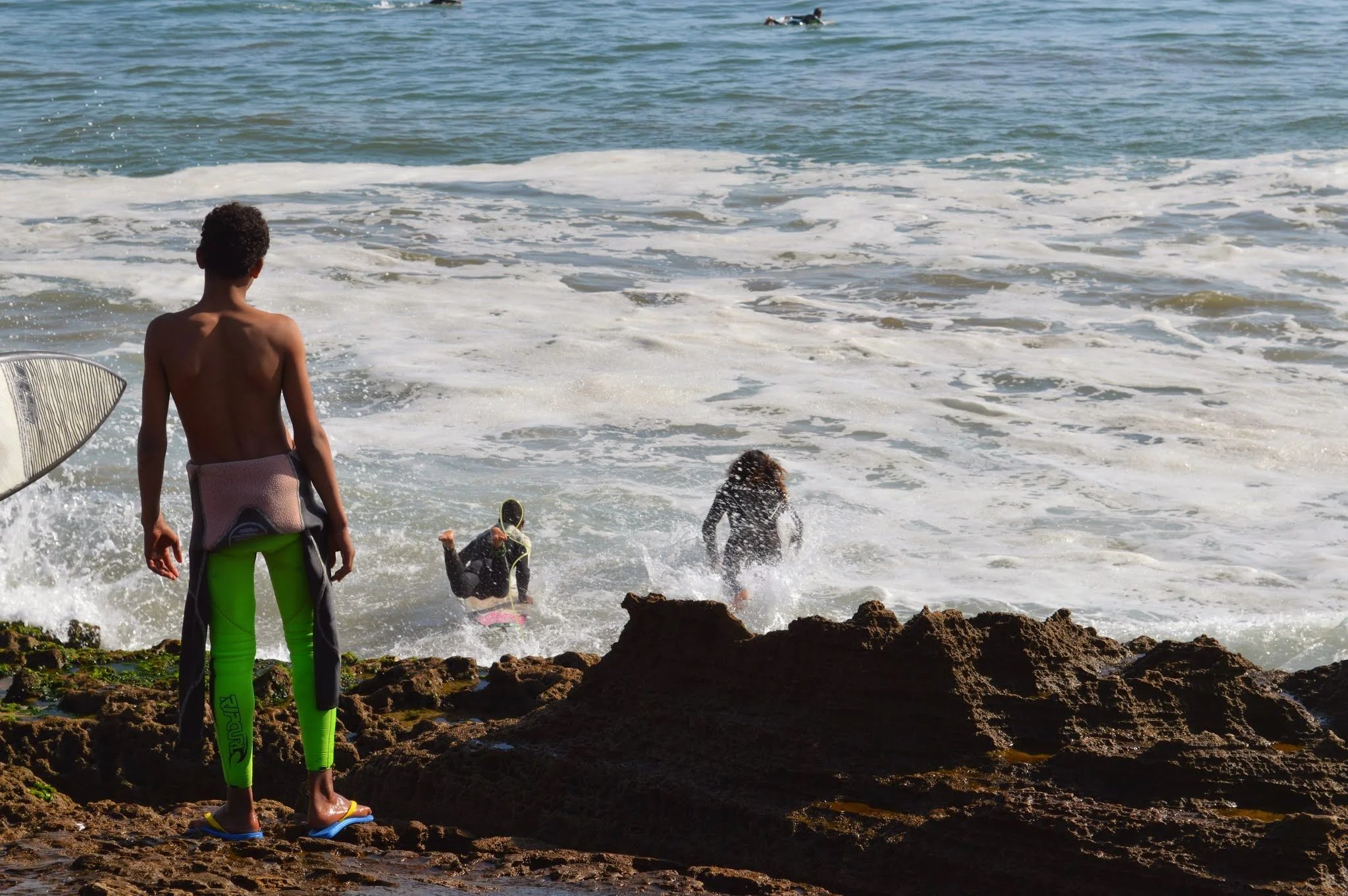 A boy standing on rocks watching children and dogs in the ocean waves at the beach.