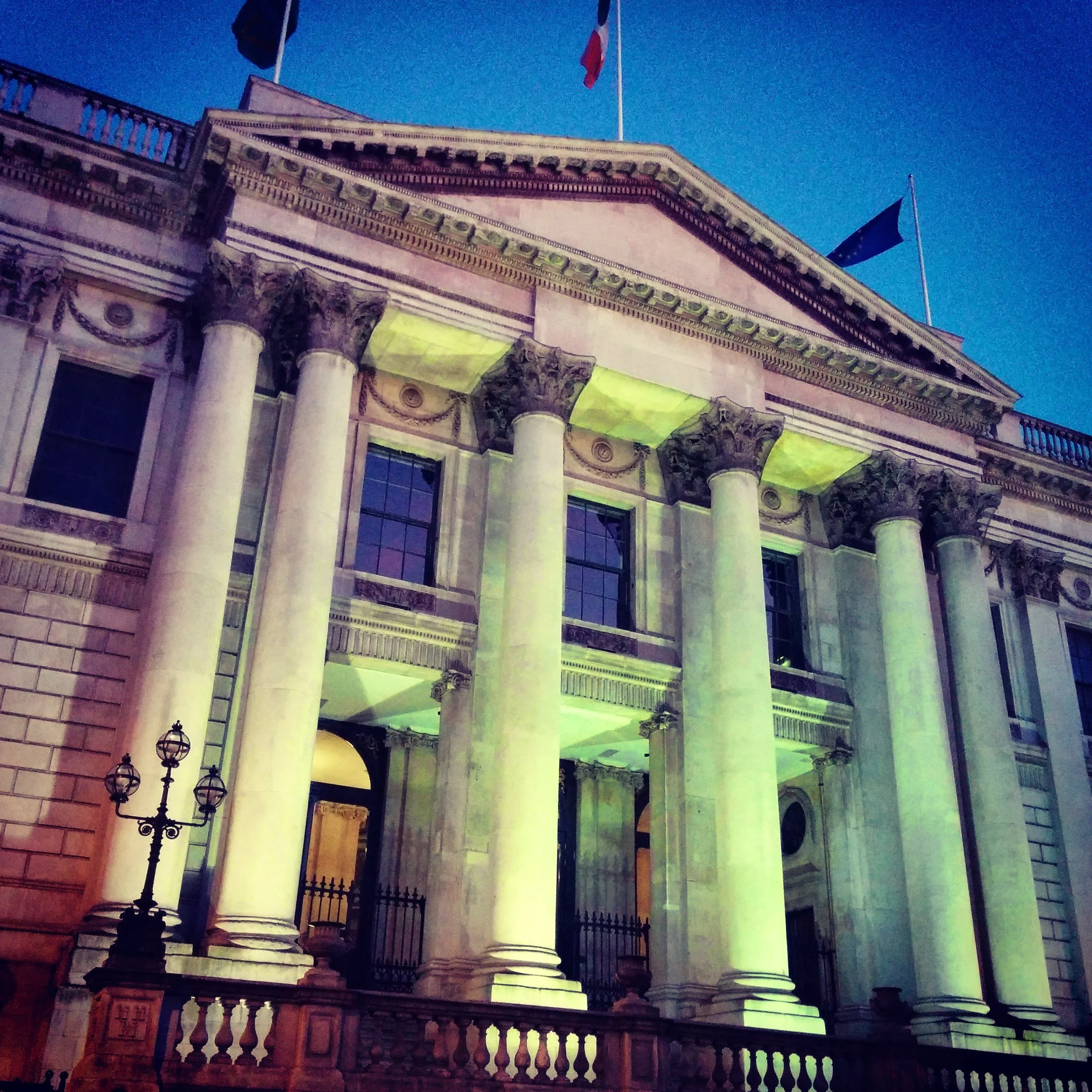 The image shows the façade of a historic building with columns, decorative carvings, and two flags flying on top, against a clear blue sky. The building is illuminated with green and yellow lighting.