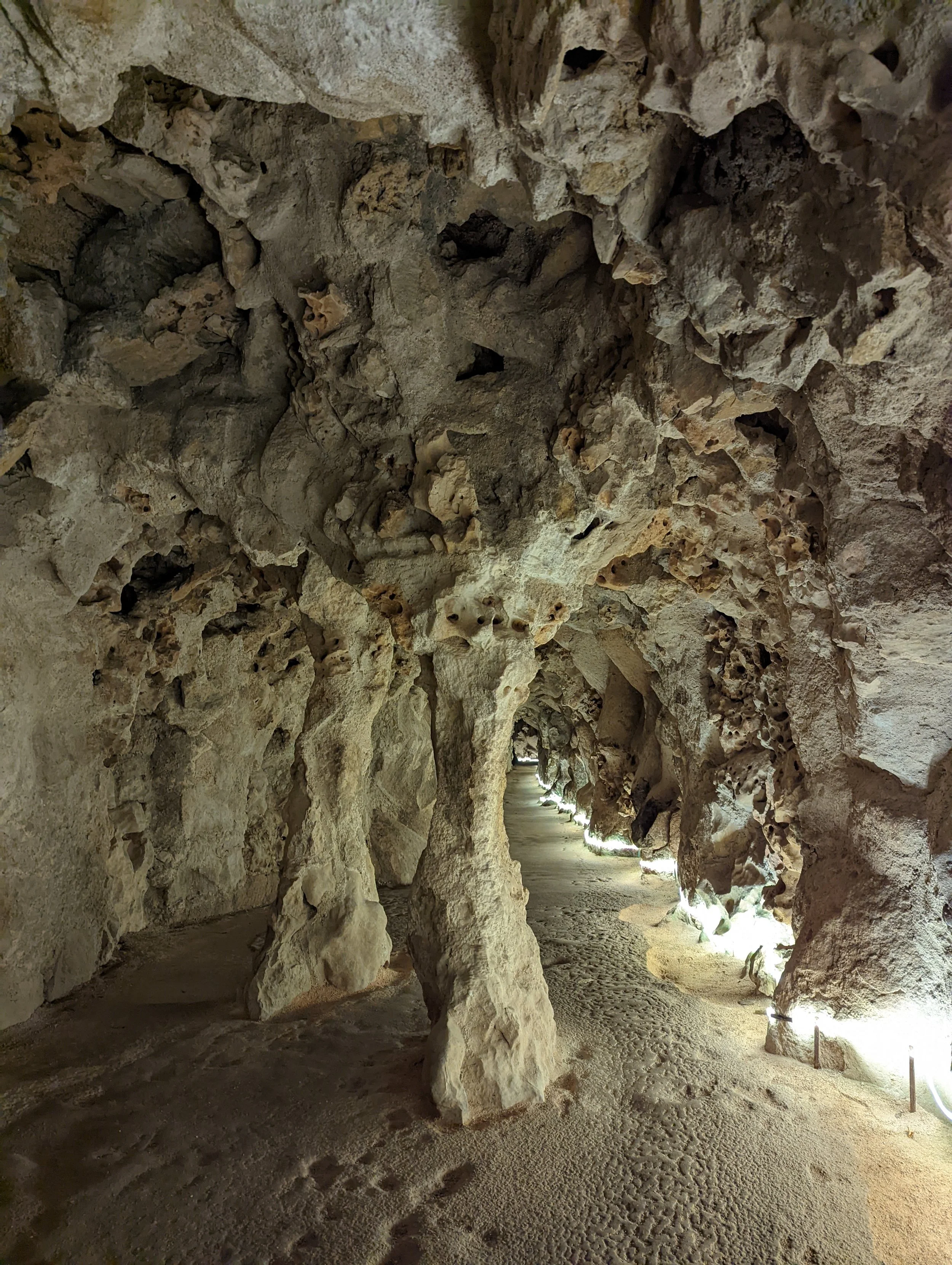 A pathway inside a stone cave with rough, textured rock walls and an uneven floor, illuminated by small lights along the side.