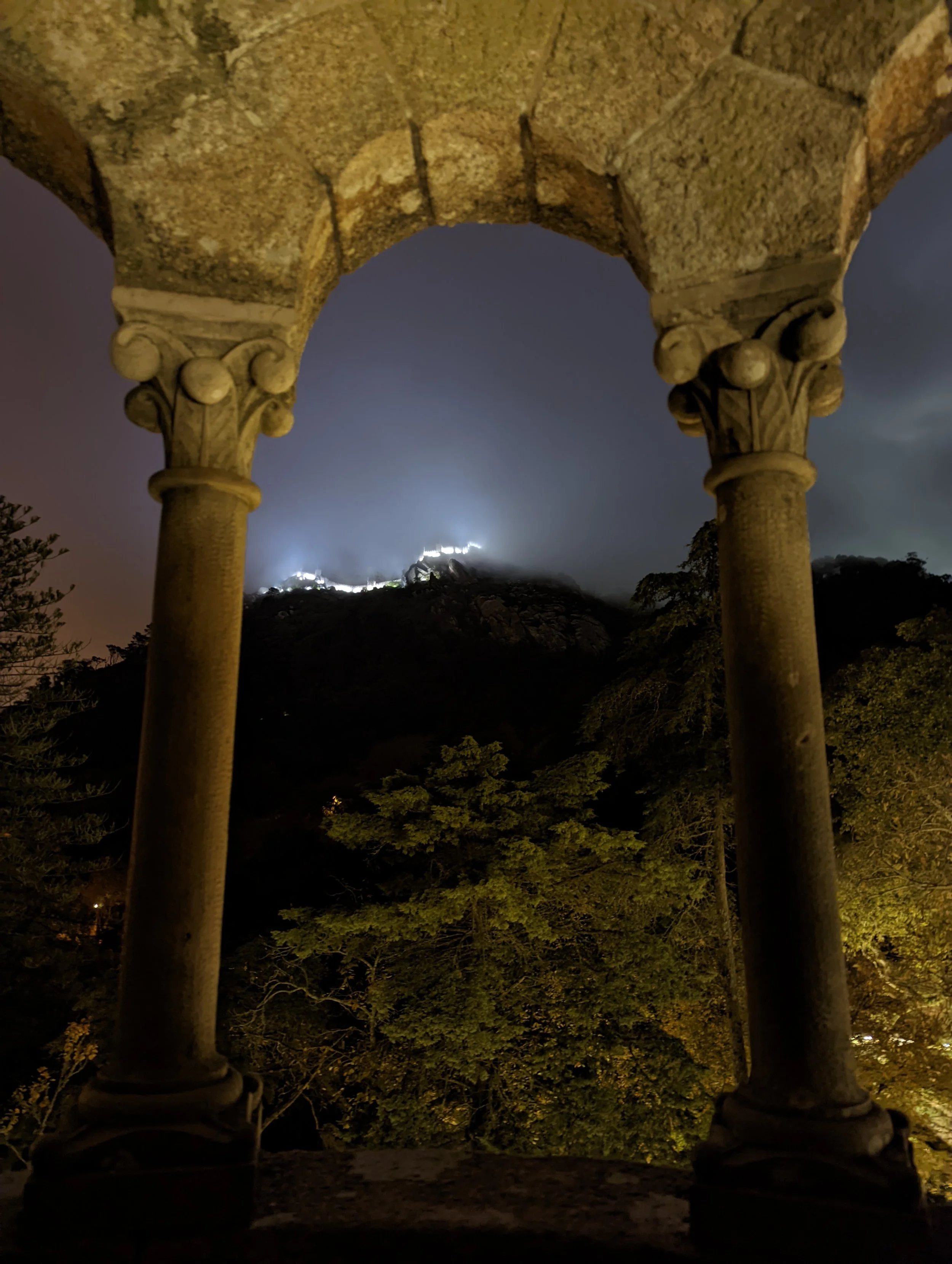 Night view through stone archway showing illuminated structure on a hill in the distance, surrounded by trees and dark sky.
