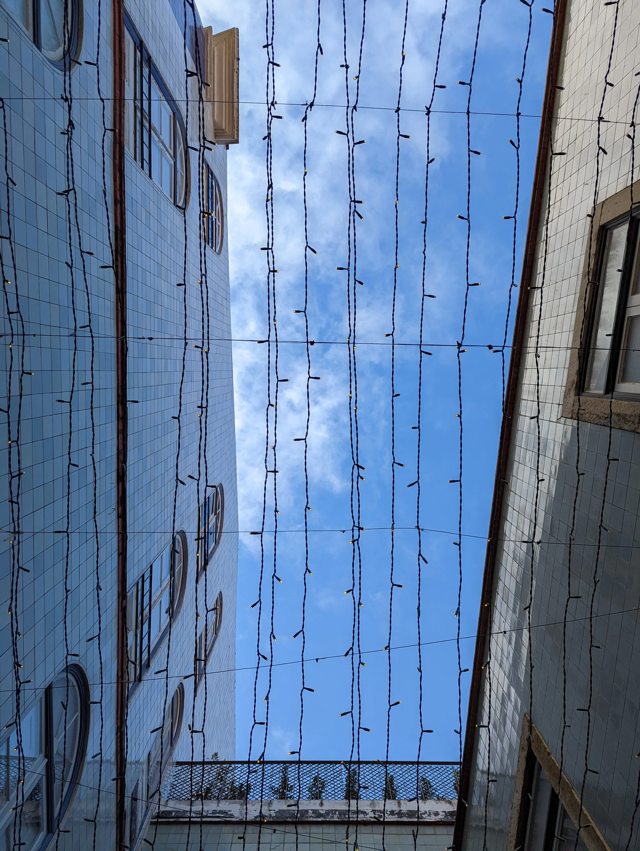 Blue sky with some clouds seen through strings of holiday lights hung between buildings with tiled exteriors.