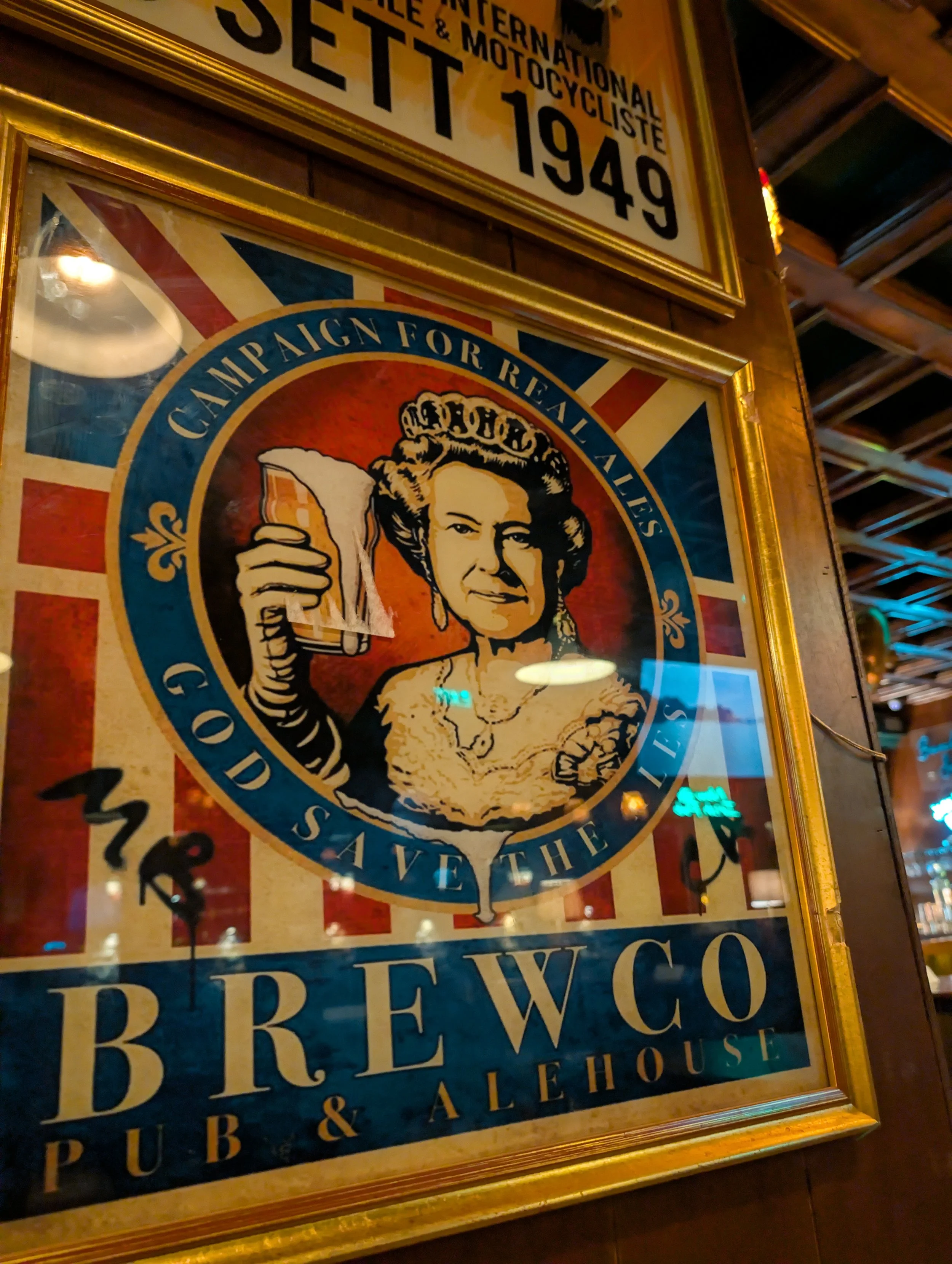Vintage framed poster of the Women's Army Auxiliary Corps with a woman holding a glass of beer, in front of a British flag and framed in gold. Partially visible text references a 1949 international motorcycle event and a 1940 law.