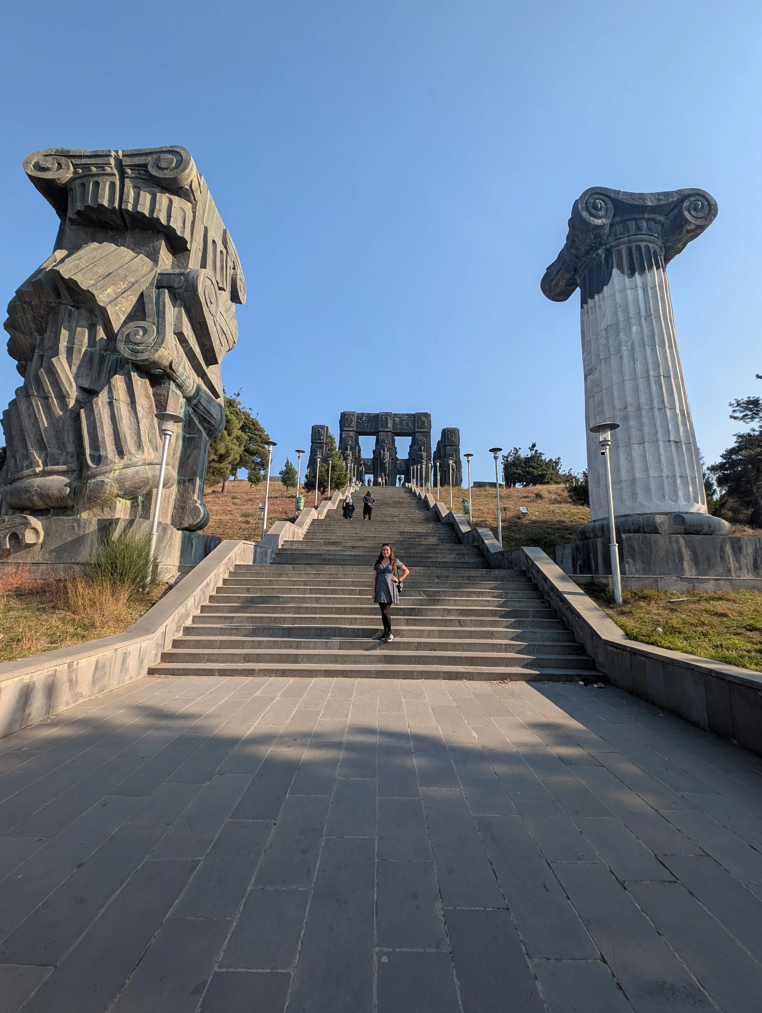 Ancient Greek ruins with large stone columns and a staircase leading up to a structure with additional columns in the background, under a clear blue sky.