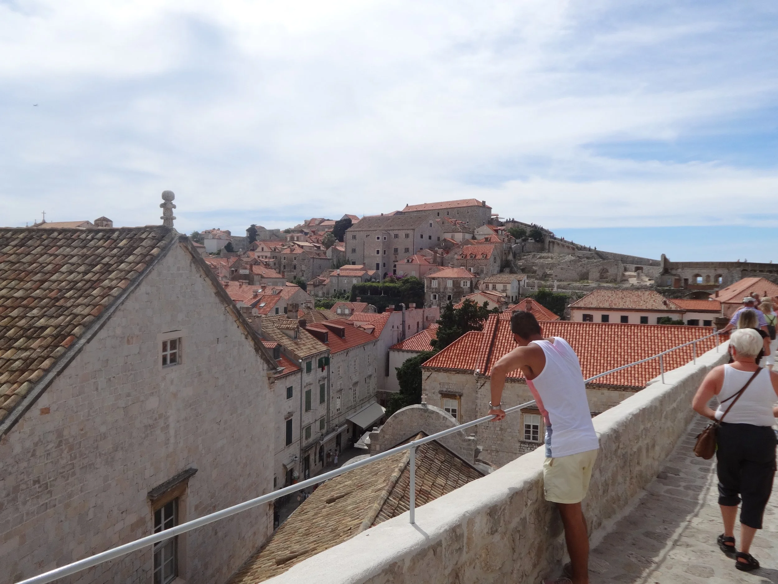 View of a historic European city with stone buildings and terracotta roofs, with people walking along a balcony or terrace overlooking the cityscape under cloudy skies.
