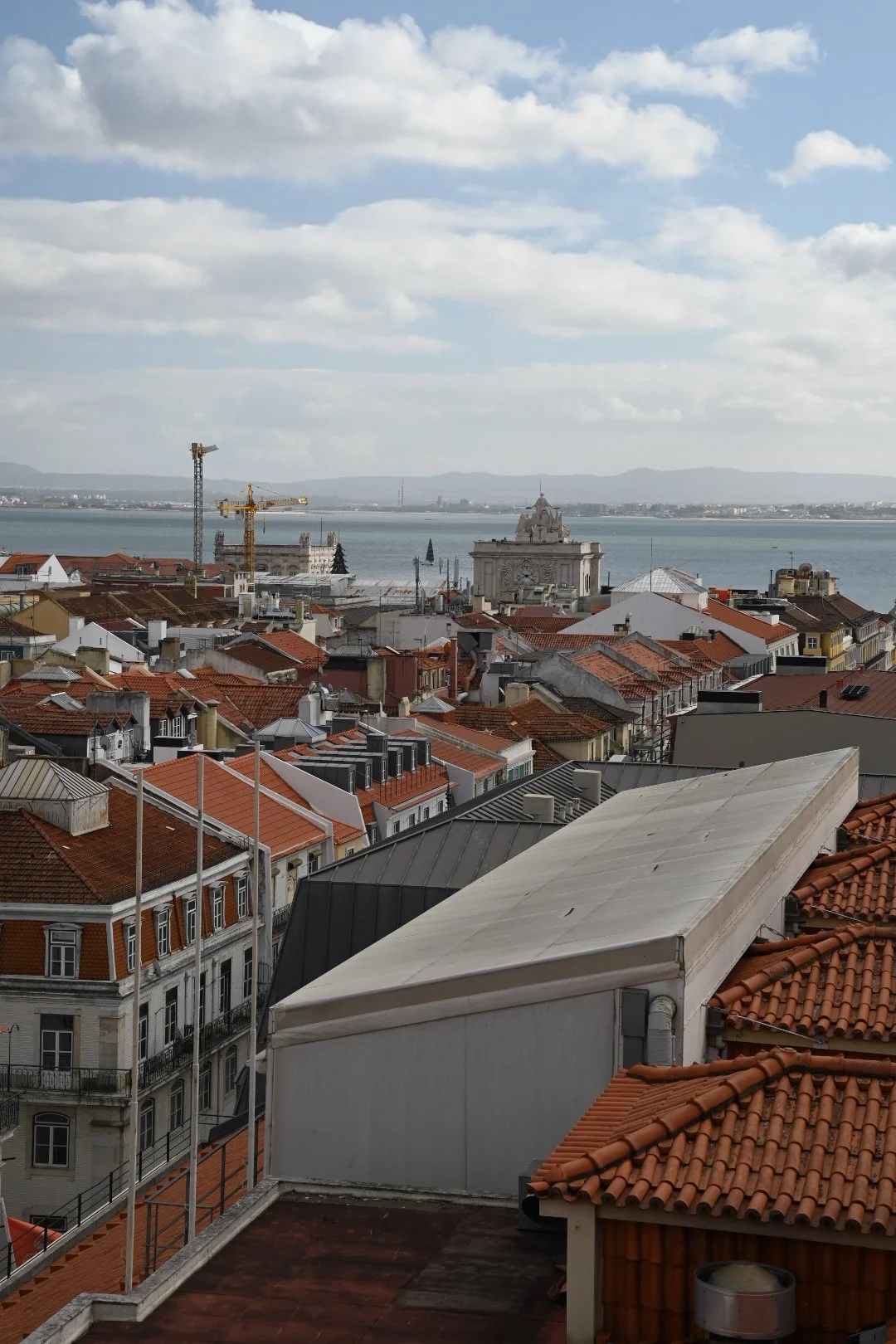Cityscape with red-tiled rooftops, a large white building with ornate architecture near the water, and construction cranes. Water and distant mountains are visible in the background under a partly cloudy sky.