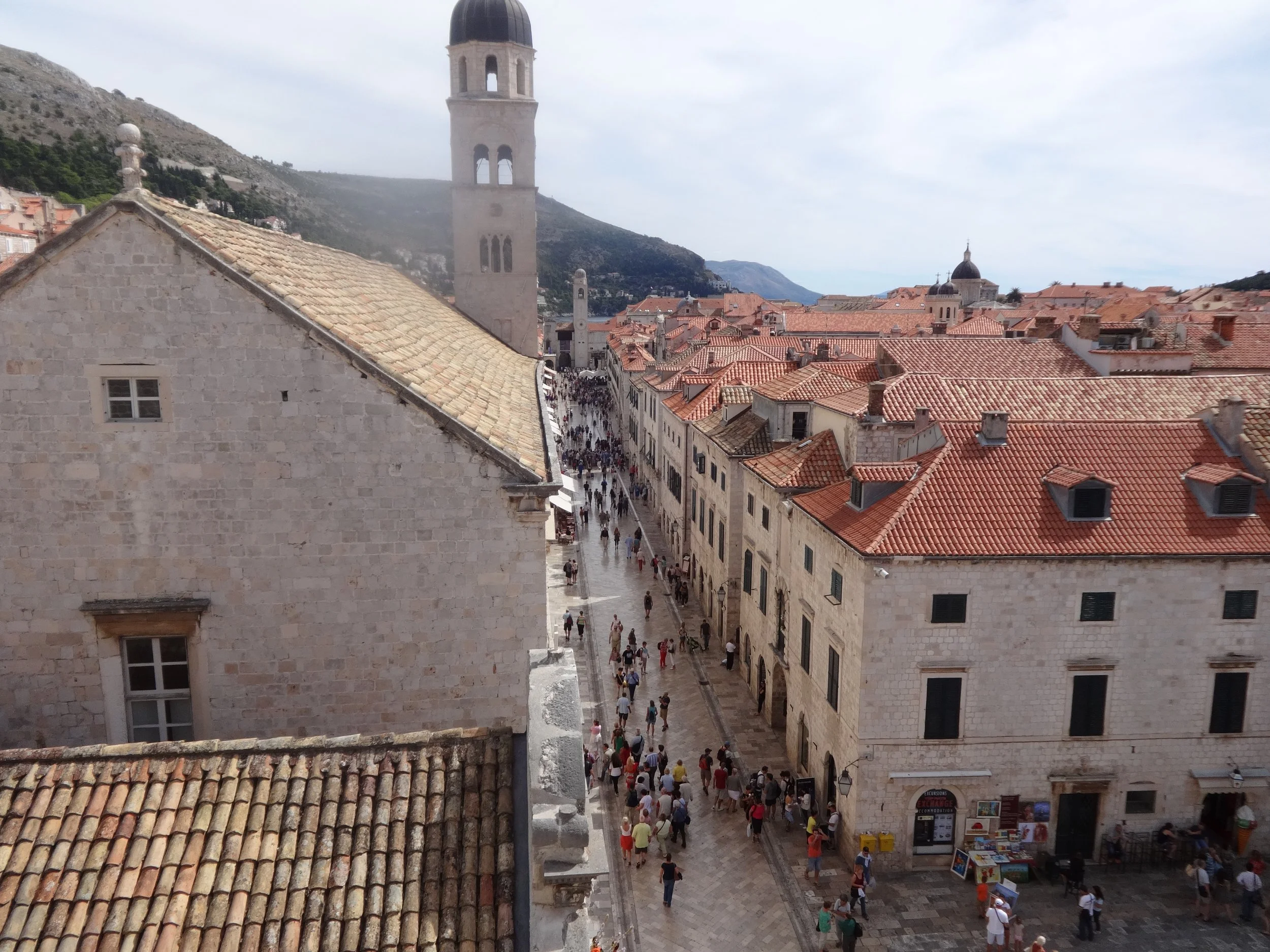 View of narrow street lined with historic stone buildings with red tile roofs, a clock tower, and a crowded street with tourists, with mountains in the background.