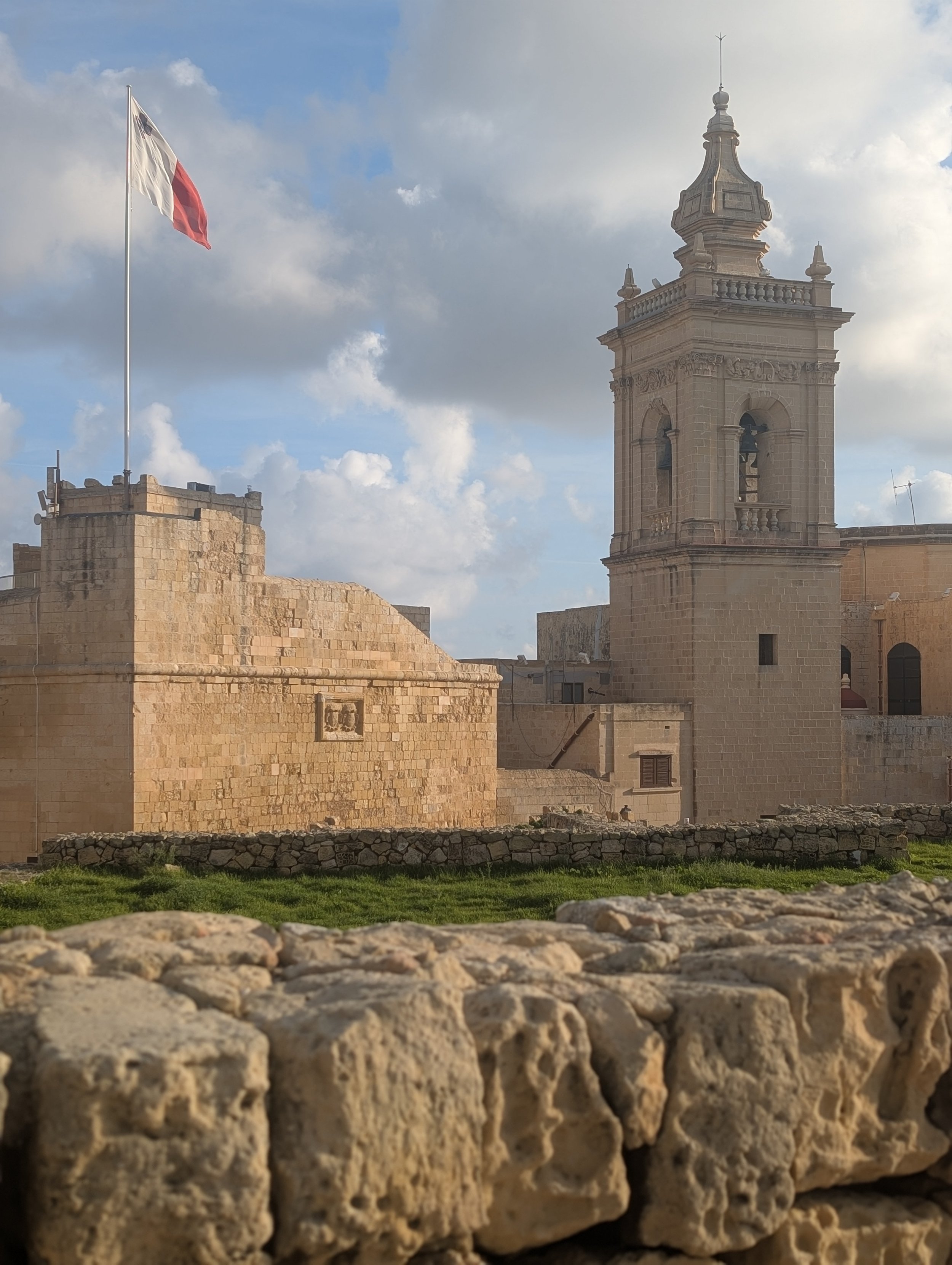 A historic stone fortress with a flagpole flying the Maltese flag and a bell tower, against a cloudy sky.
