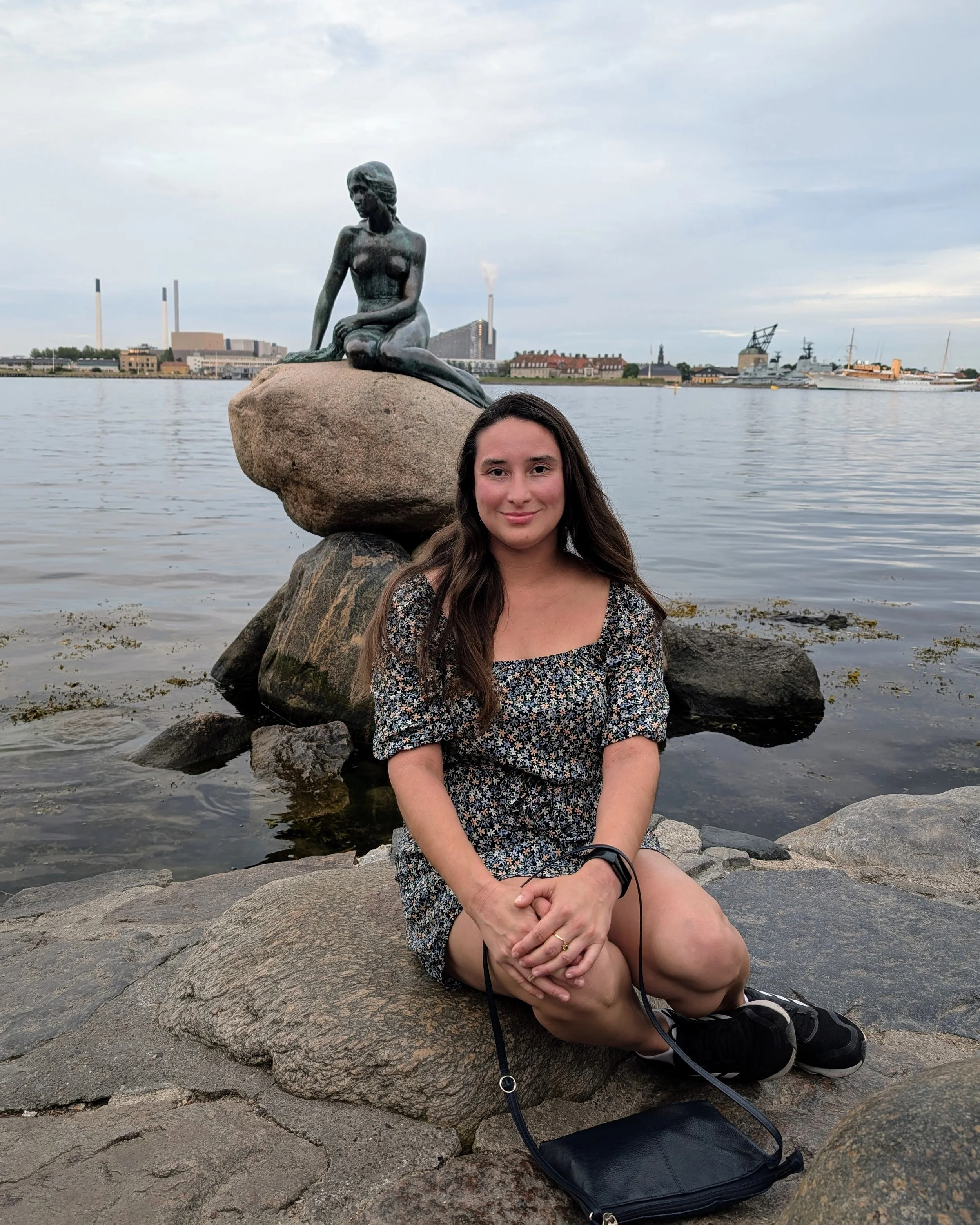 A woman with long brown hair sitting on rocks by the water, with a statue of a mermaid on a large rock behind her. The background includes industrial buildings, ships, and a cloudy sky.