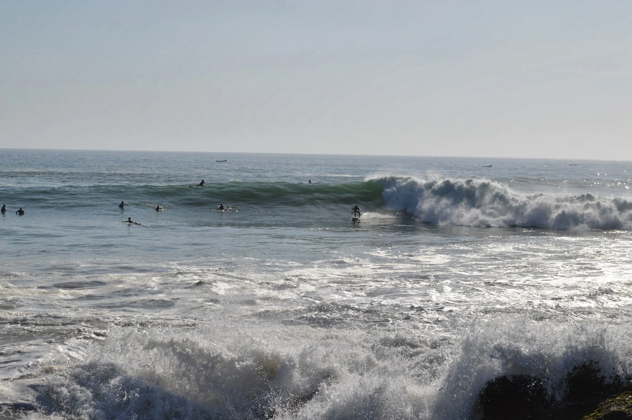A beach scene with waves, surfers in wetsuits, and boats on the horizon under a clear sky.
