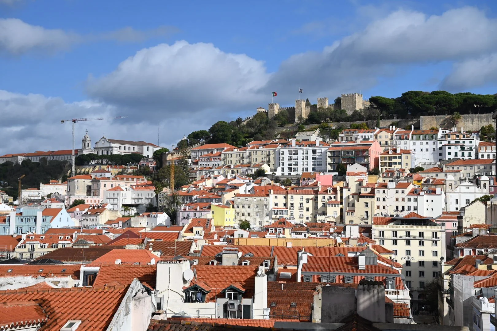 A hillside view of a city with colorful buildings and red-tile roofs, featuring a castle on top of the hill in the background and a bright blue sky with clouds.