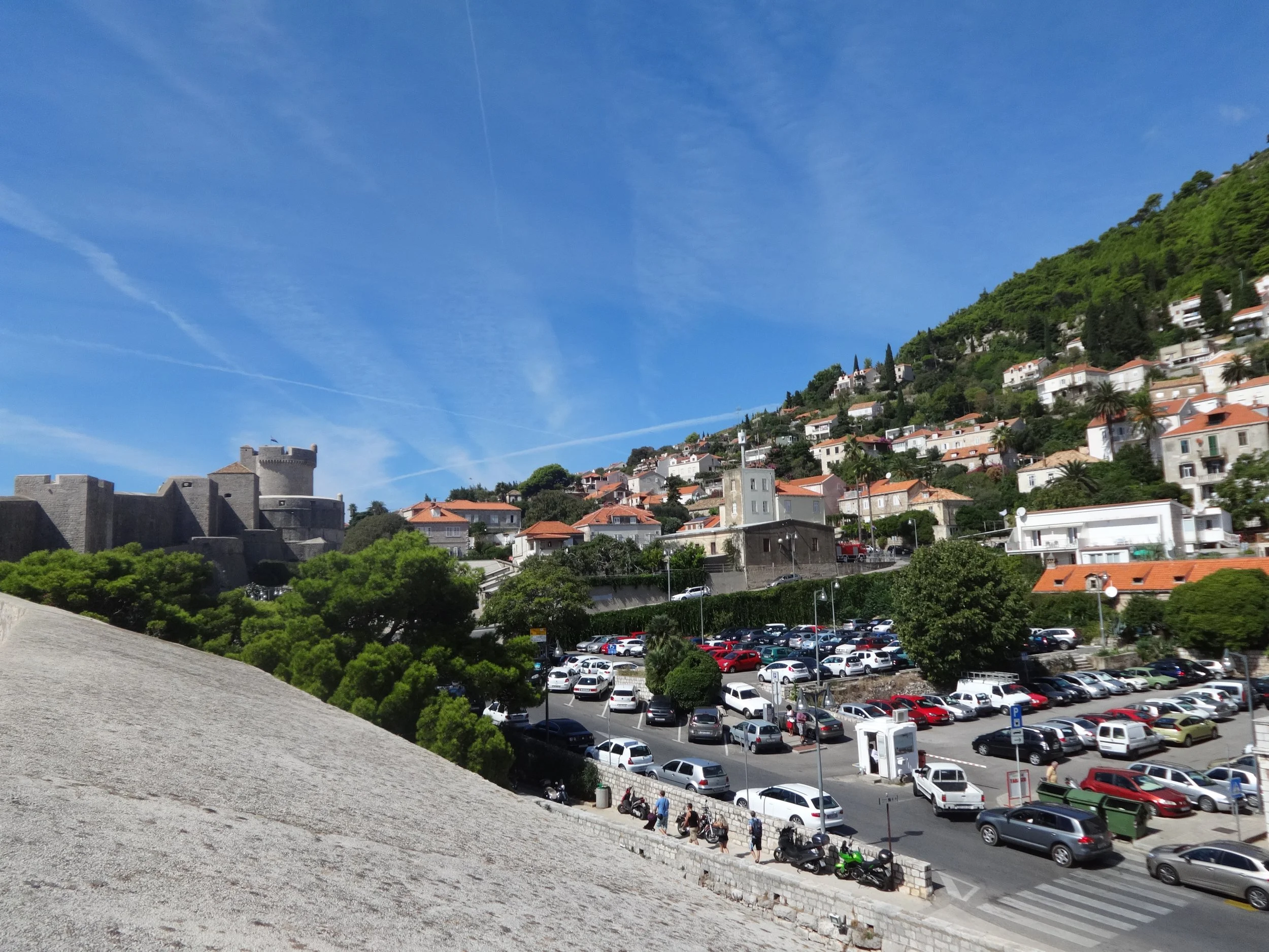 View of a coastal town hillside with white buildings and red rooftops, a castle or fortress wall in the foreground, and a parking lot filled with white and silver cars, under a clear blue sky with thin clouds.