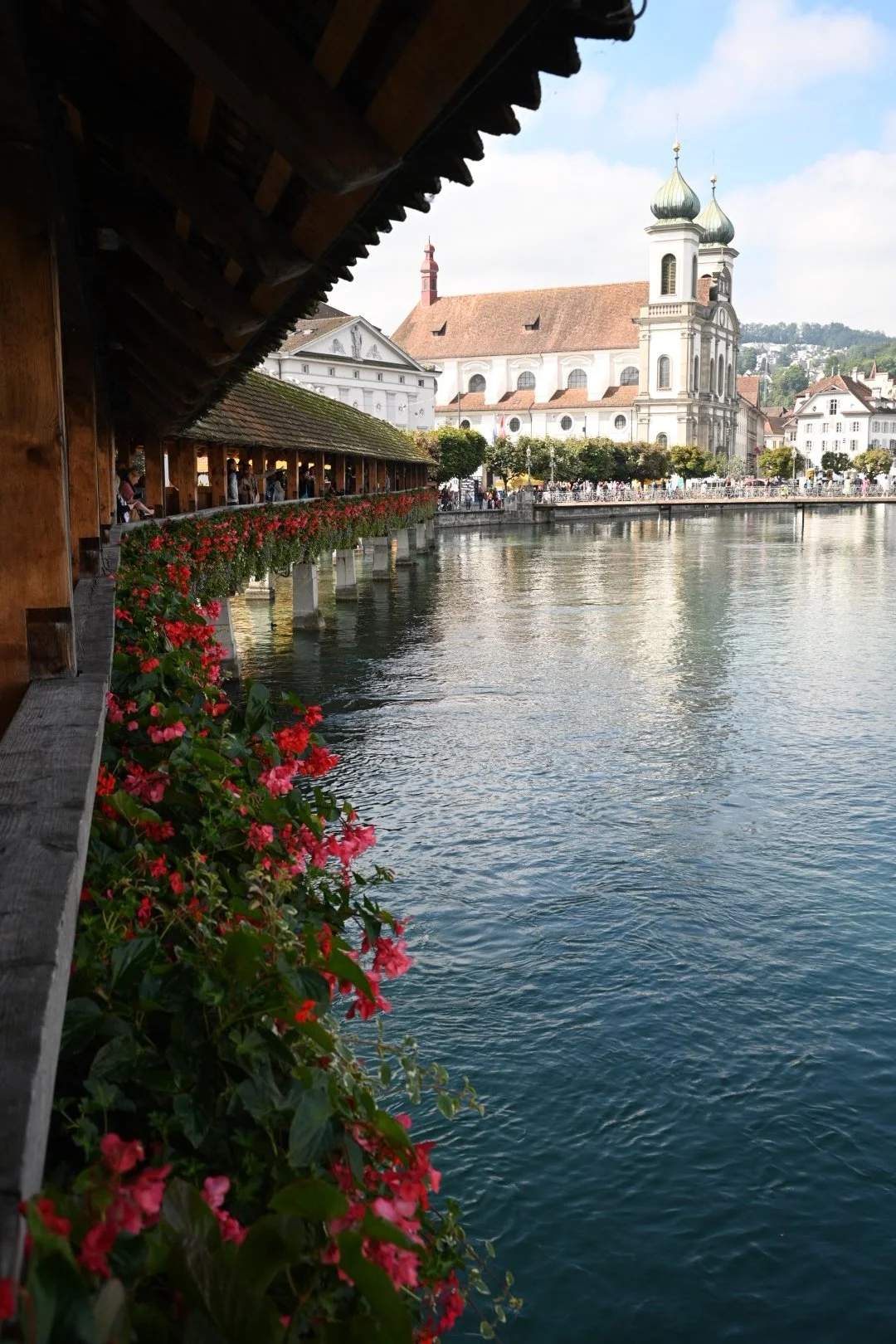 A scenic view of a lakeside town with a church in the background, a wooden covered bridge with red flowers hanging along its side, and a calm body of water reflecting the buildings.