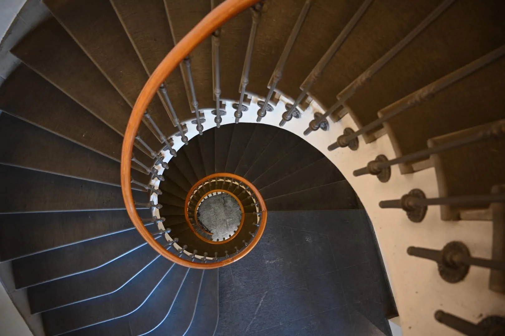 Top-down view of a spiral staircase with dark stair treads, metal balusters, and wooden handrails, creating a vortex pattern.