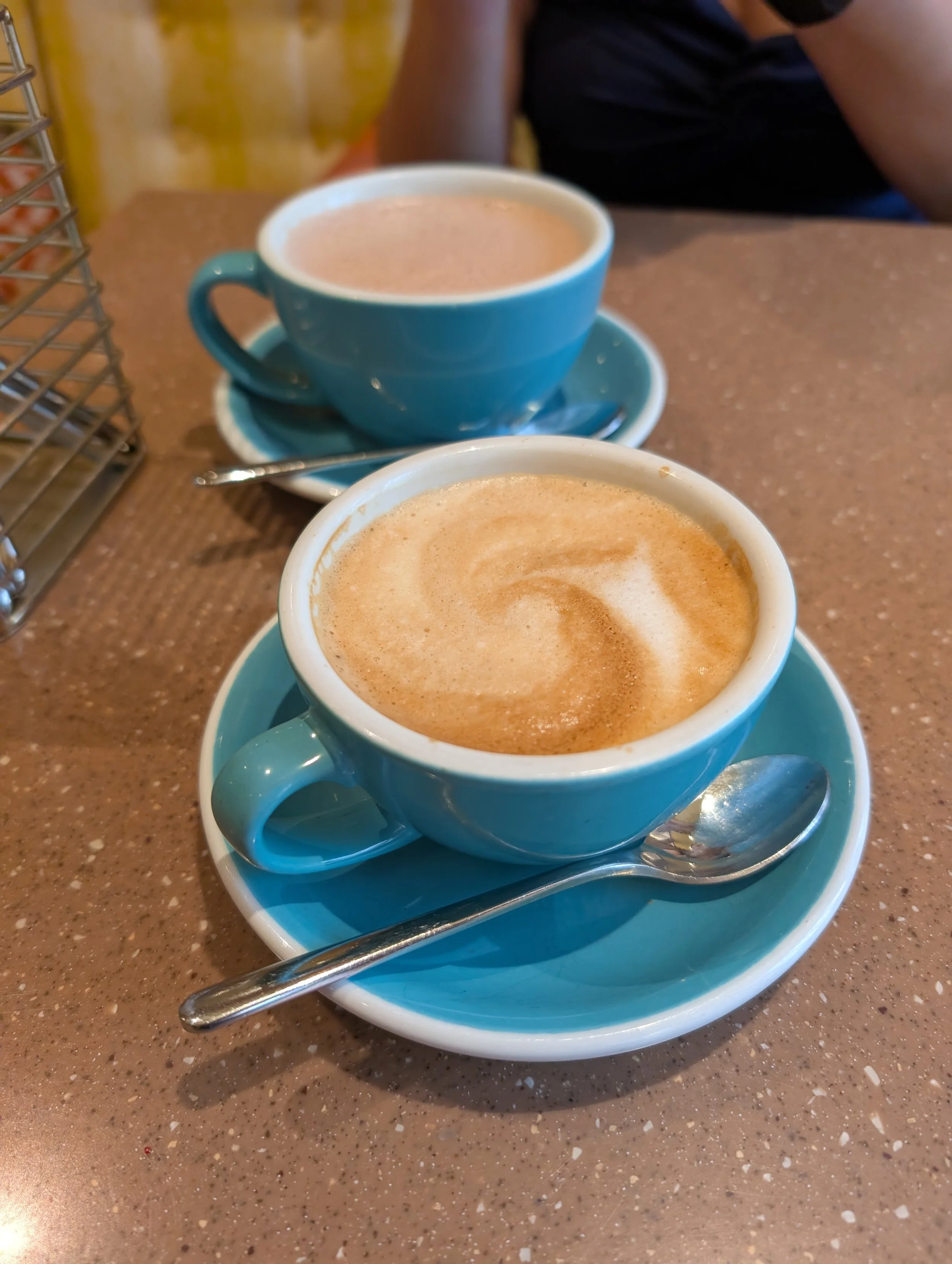 Two cups of coffee with frothy milk served on blue cups and saucers on a brown speckled table, with spoons resting on the saucers.