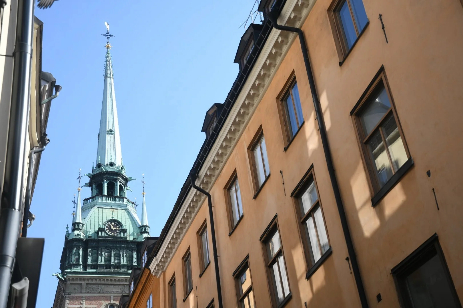 Tall clock tower with green roof and spire behind yellow residential buildings with multiple windows under a clear blue sky.