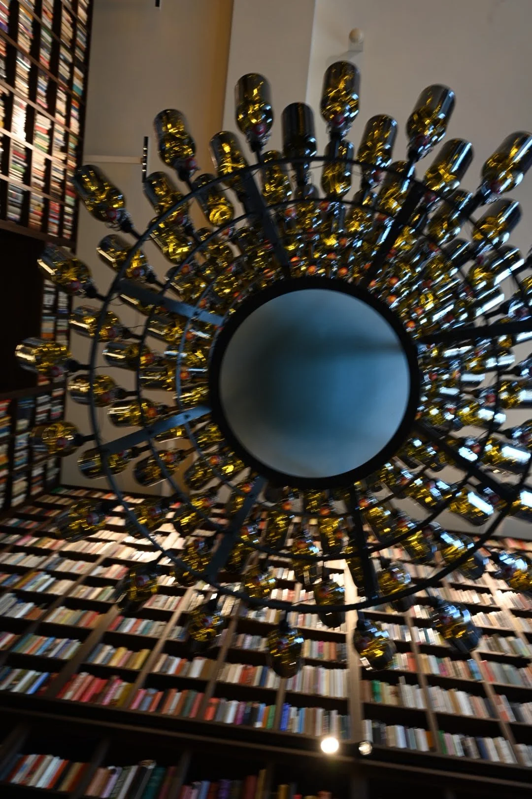 A view of a circular chandelier with numerous golden bulbs hanging from the ceiling, with a large bookshelff filled with books in the background.