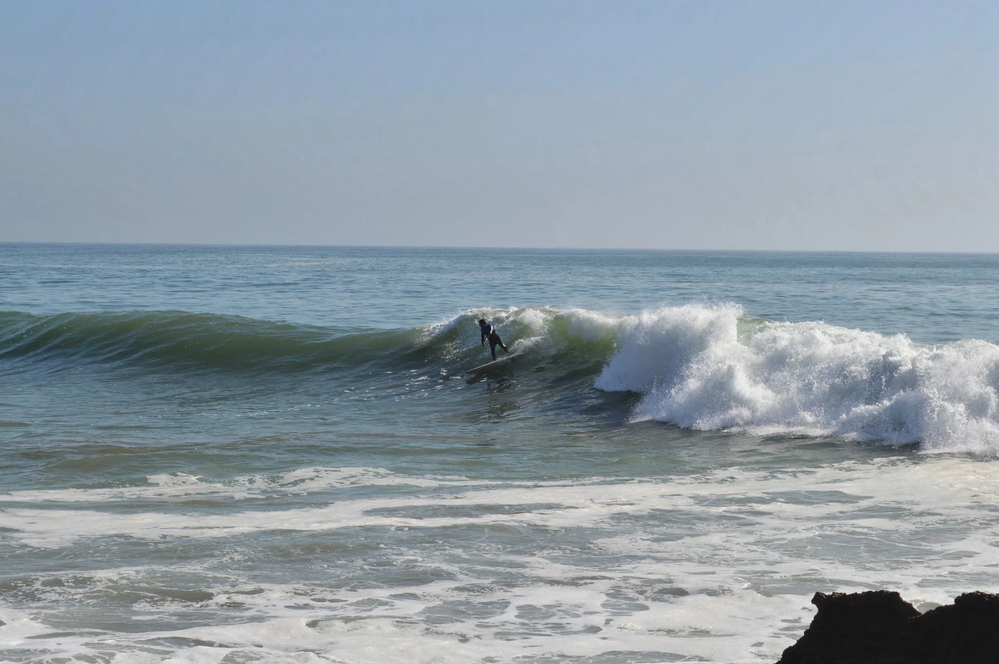 A person surfing on a wave in the ocean near the shoreline with rocks visible in the foreground.