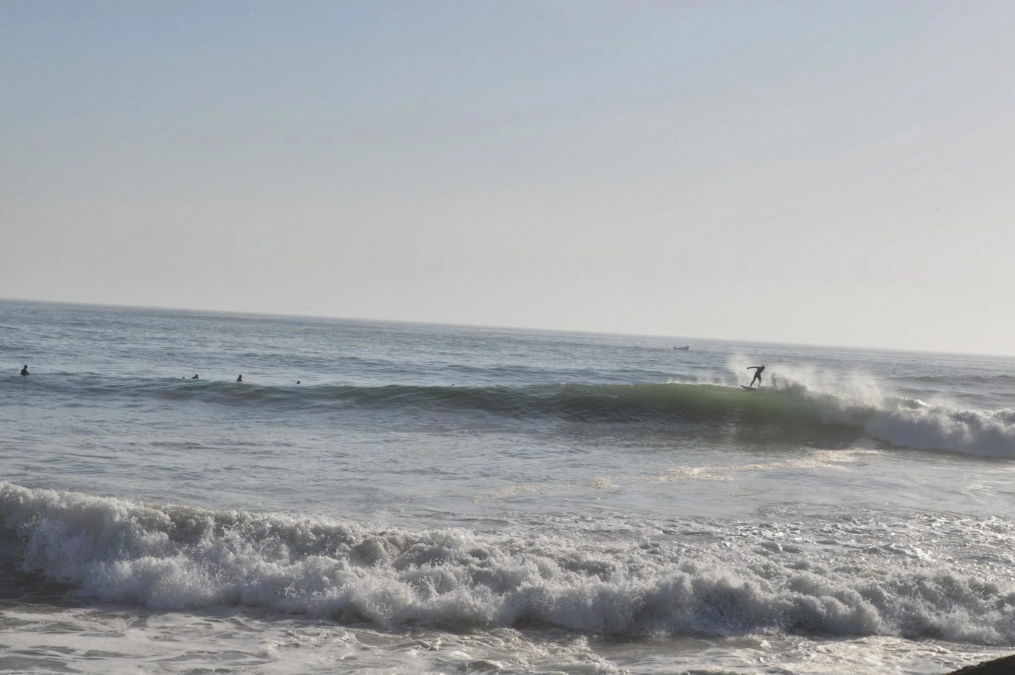 Surfer riding a wave near the shoreline with several people swimming and watching nearby, ocean scene with a boat in the distant background.