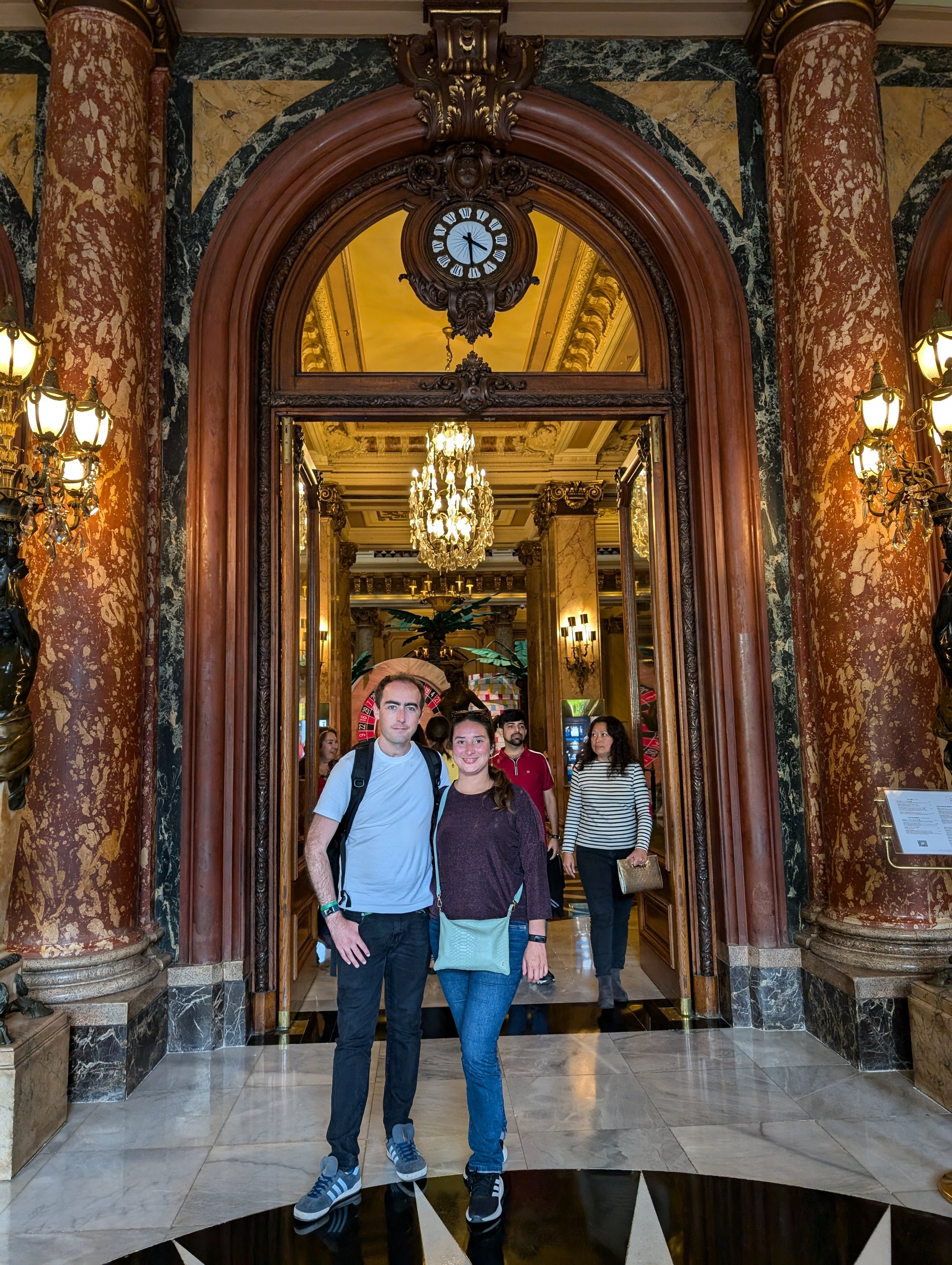 A group of people standing in front of an ornate, antique mirror in a luxurious, gold and marble decorated interior. The mirror is framed with dark wood and gold accents, and a clock is mounted above it showing just past 3:00. The ceiling and chandel