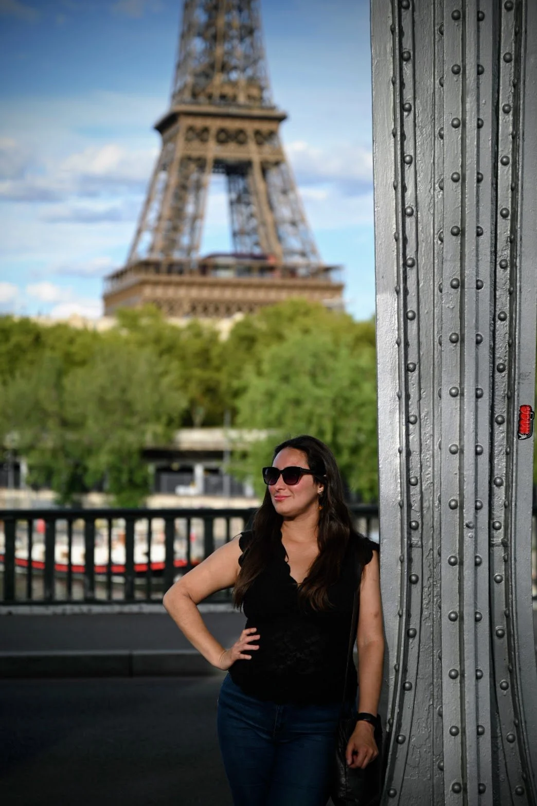 A woman with long dark hair, wearing sunglasses, a black sleeveless top, and jeans, poses with her hand on her hip near a metal structure on a bridge, with the Eiffel Tower and green trees in the background.