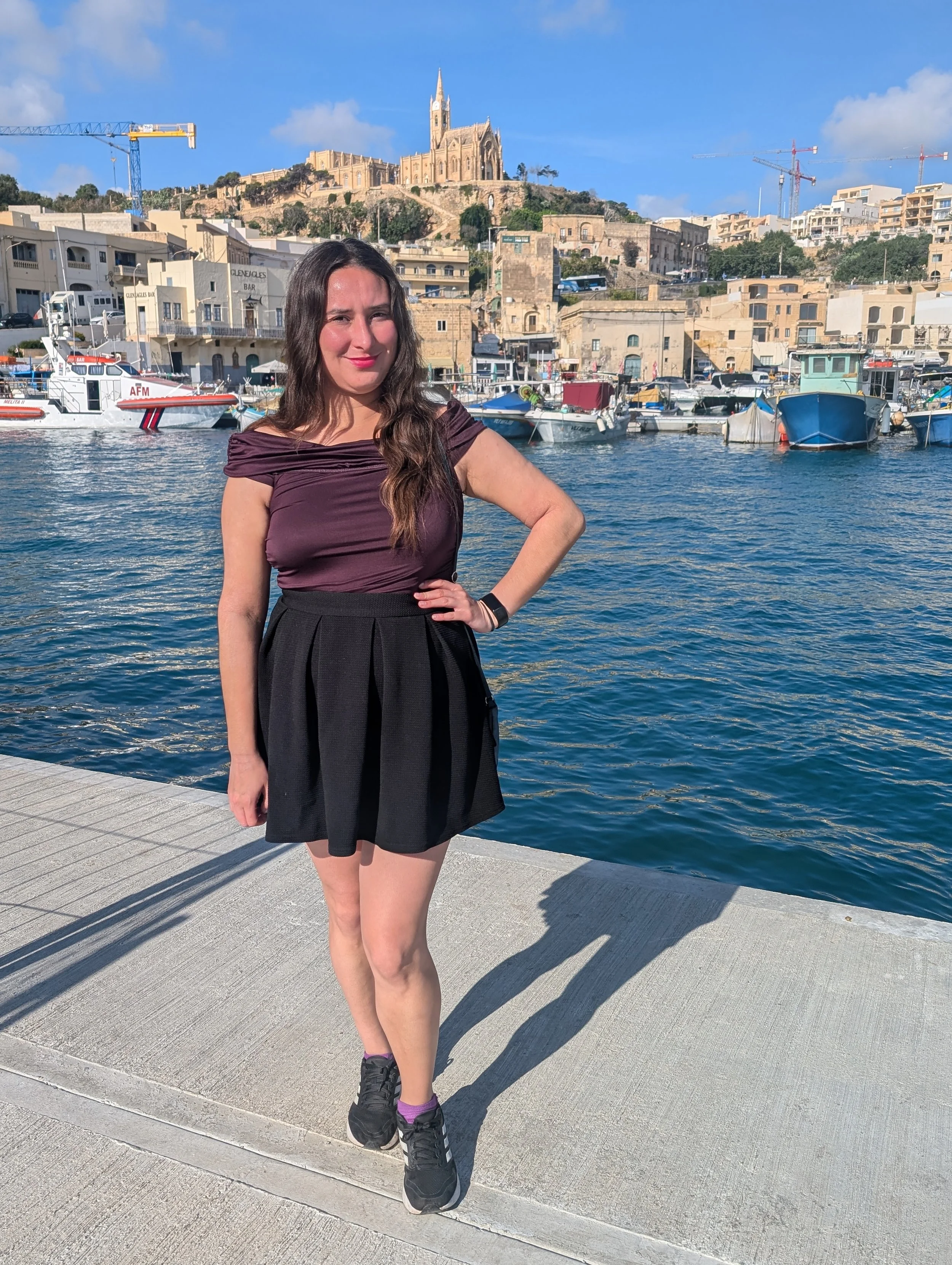 A woman with long brown hair in a purple top and black skirt standing on a waterfront promenade with boats docked behind her. A hillside with buildings, trees, and a church with a tall spire is visible in the background on a sunny day.