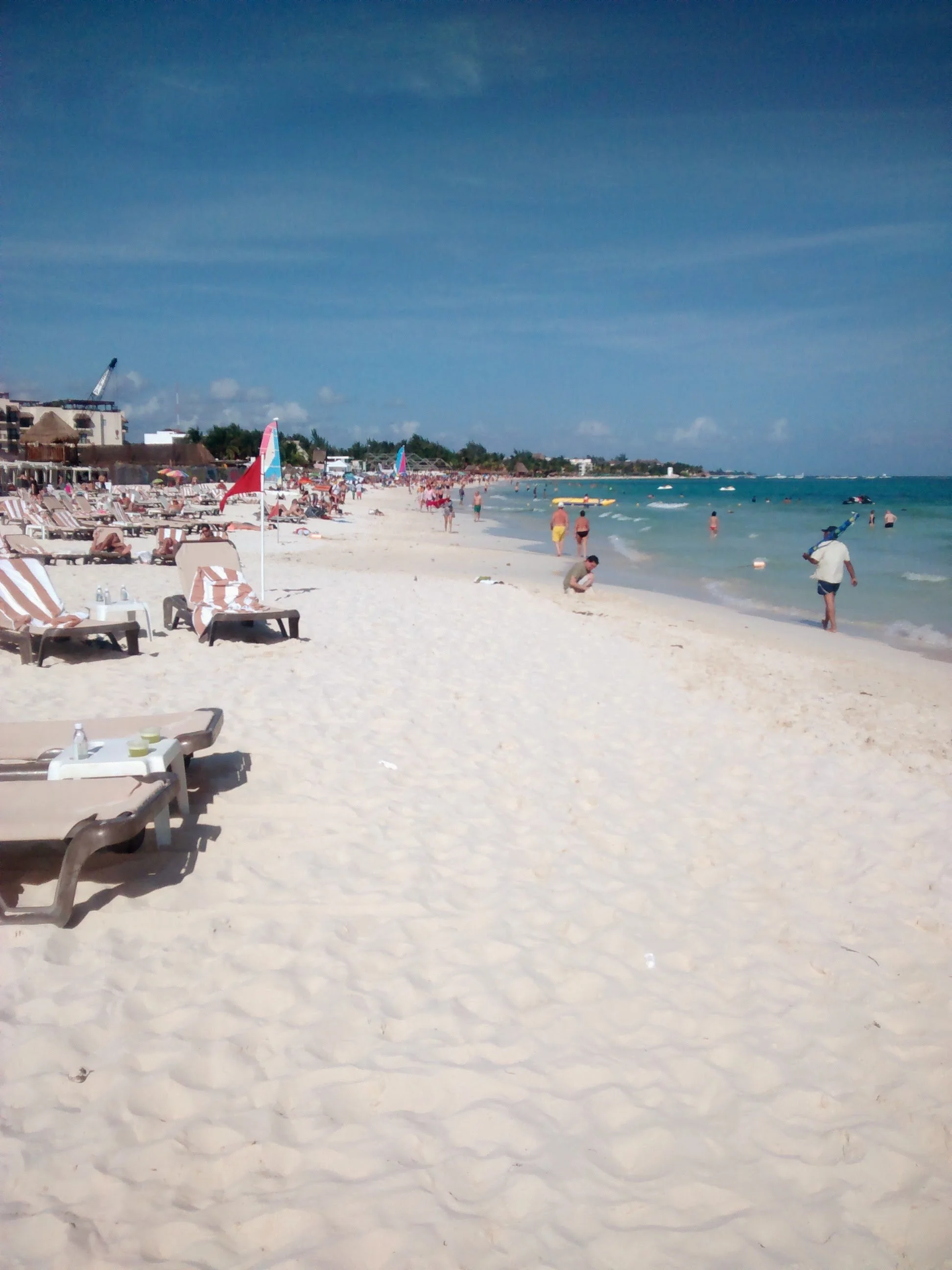 Beach with white sand, lounge chairs, umbrellas, and people swimming and walking along the shoreline, with blue sky and buildings in the background.