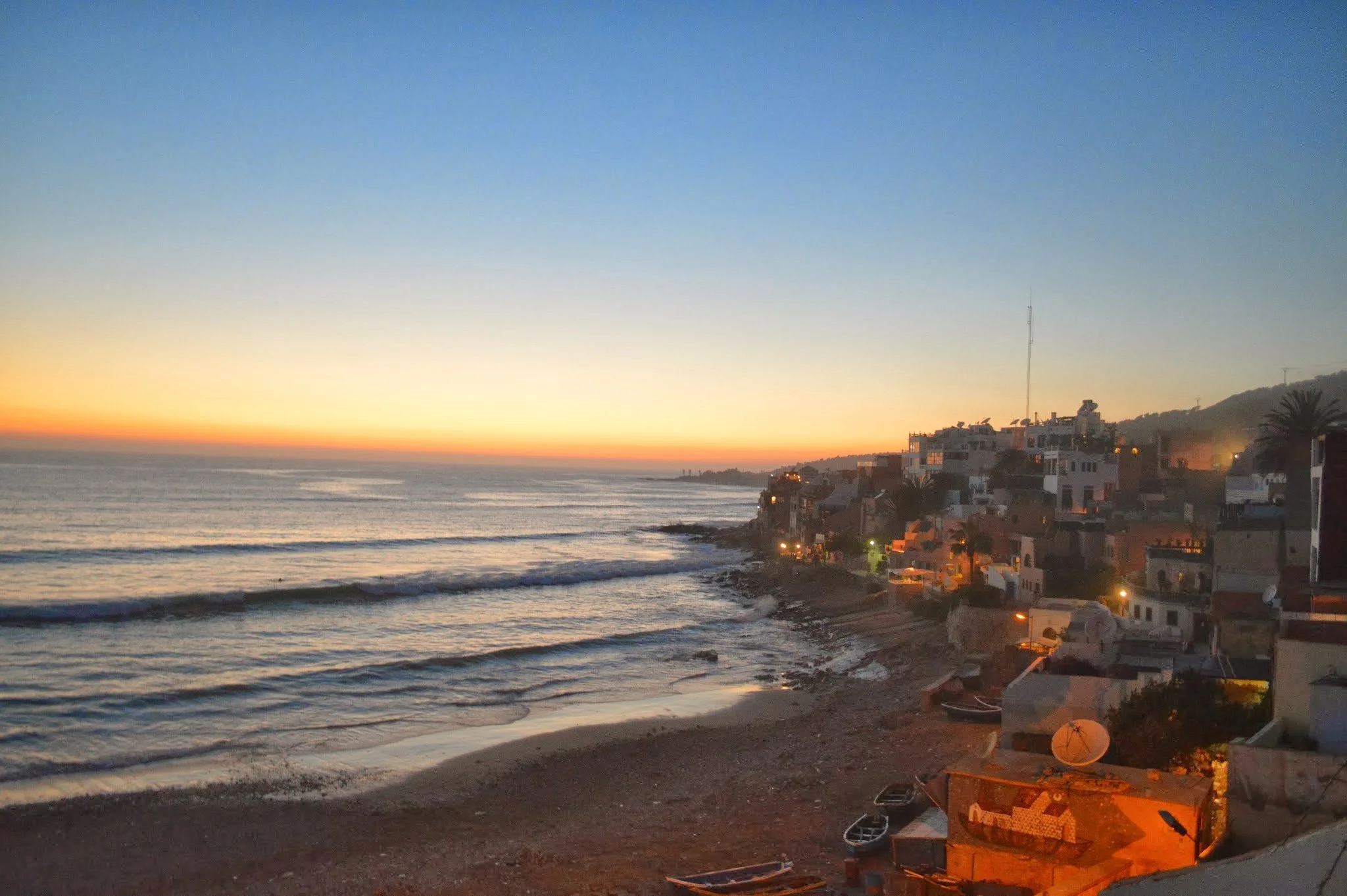 Sunset over the ocean with a shoreline and coastal houses illuminated by warm lights.