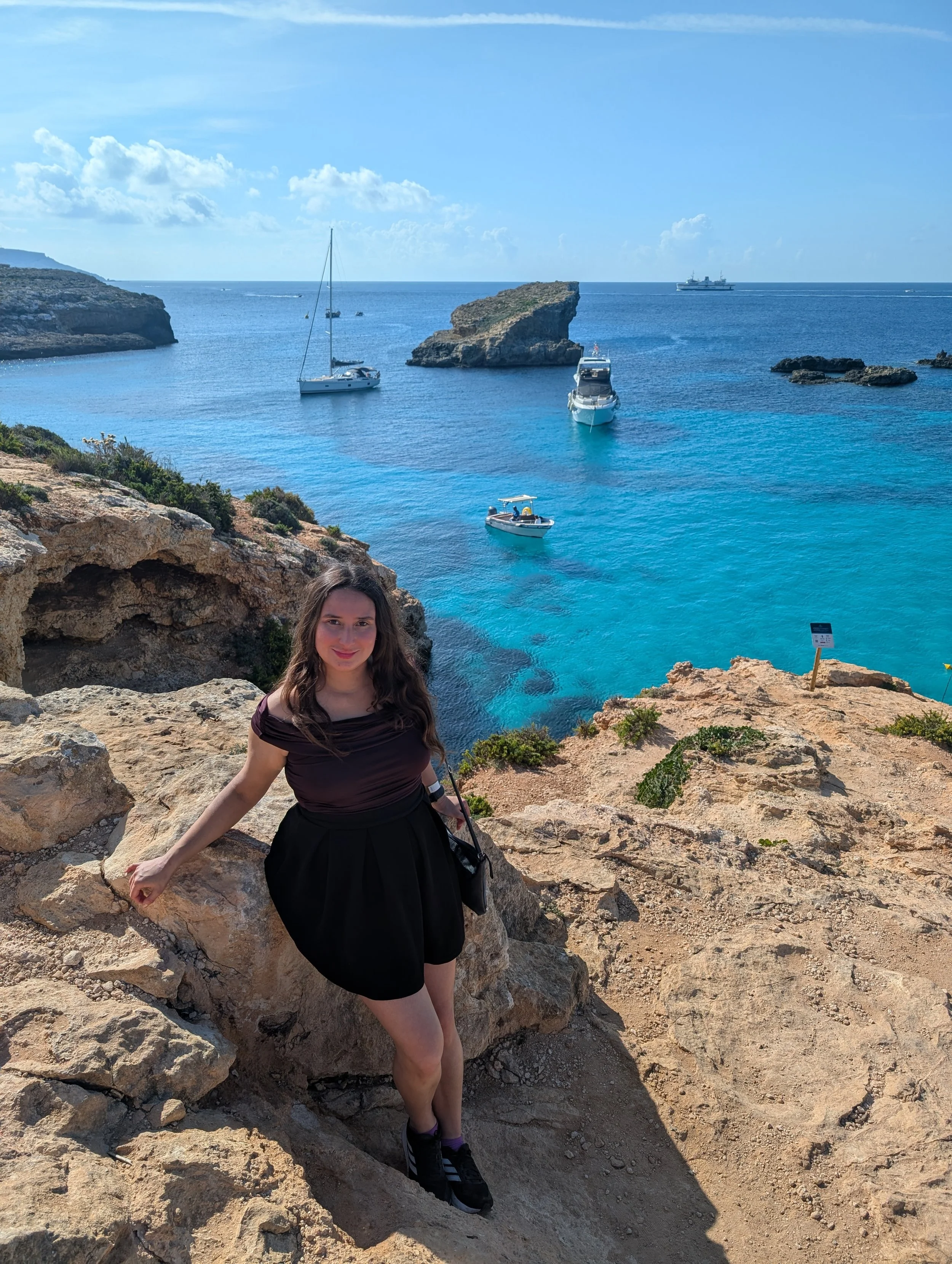 A woman with long brown hair and a black skirt posing on rocky terrain by the ocean, with boats and clear blue water in the background on a sunny day.