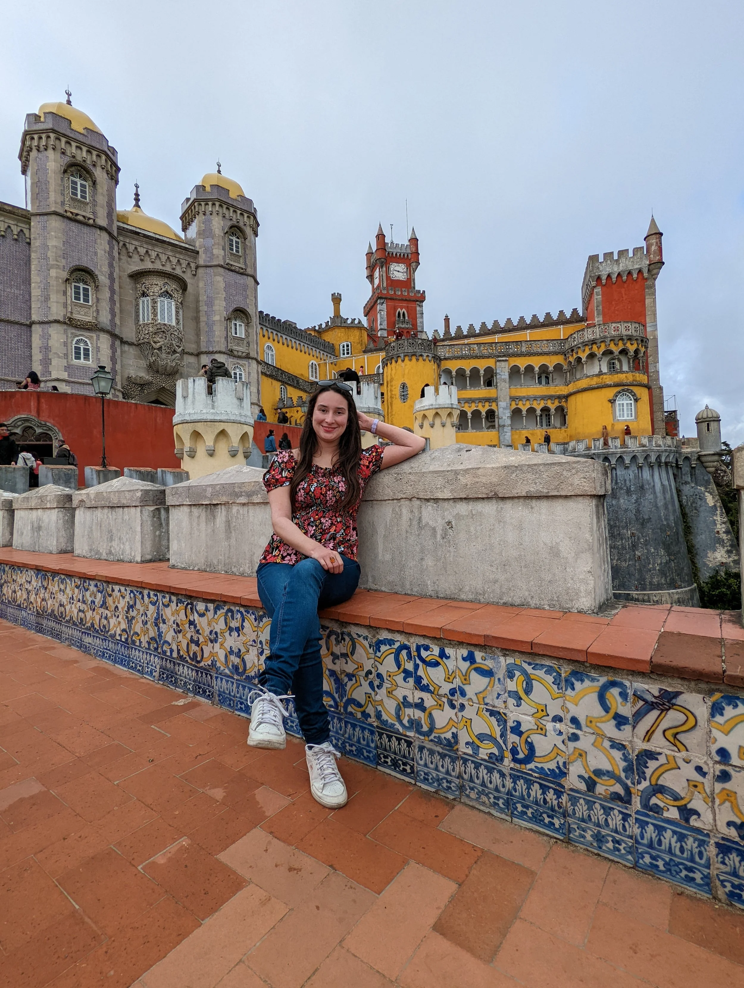A woman wearing a floral top and jeans sitting on a decorative tiled wall in front of colorful historic castle with turrets, towers, and a clock, under overcast skies.