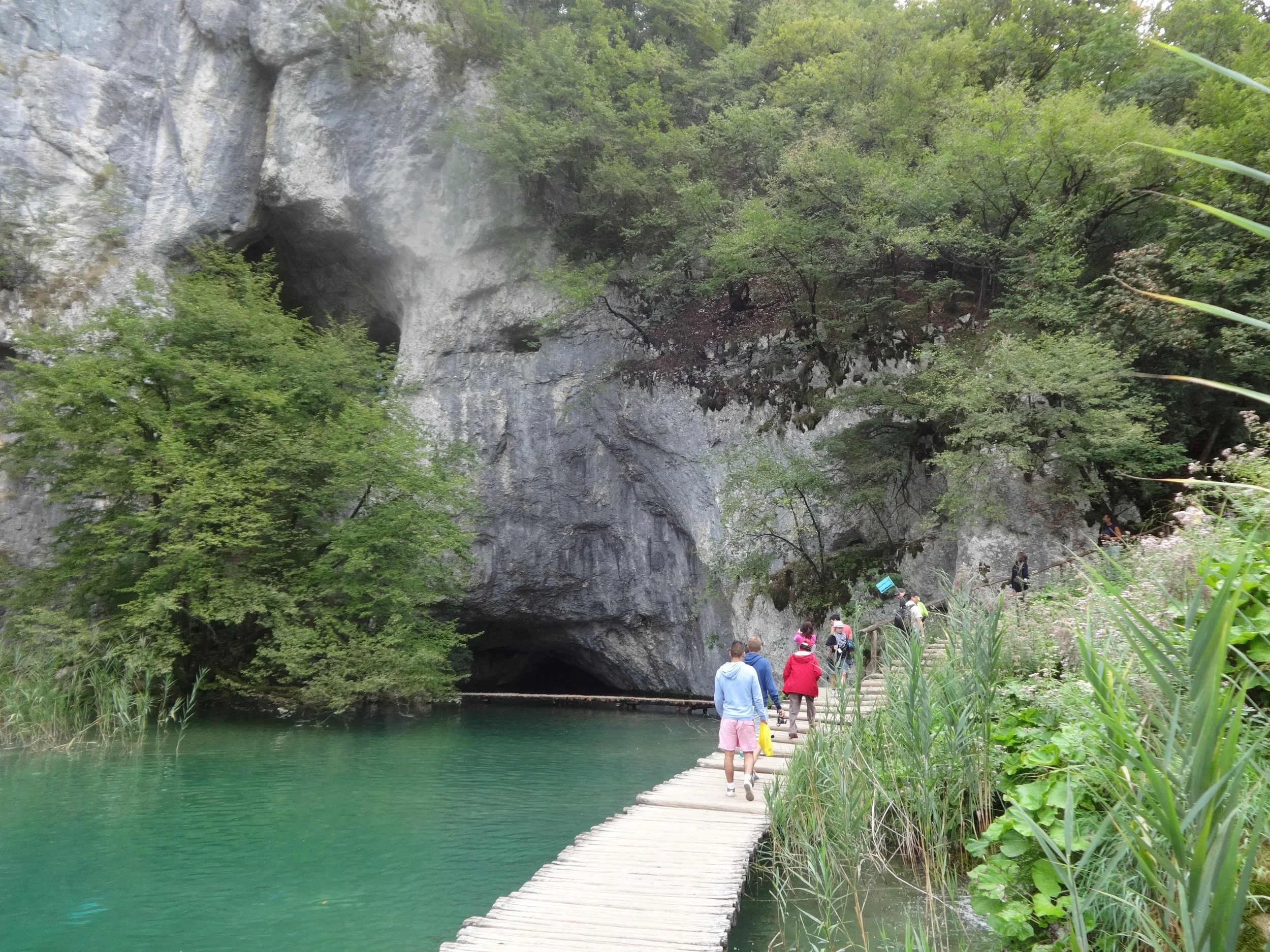 People walking on a narrow wooden path beside a green lake, surrounded by lush trees and a rocky mountain with caves and mist in the background.
