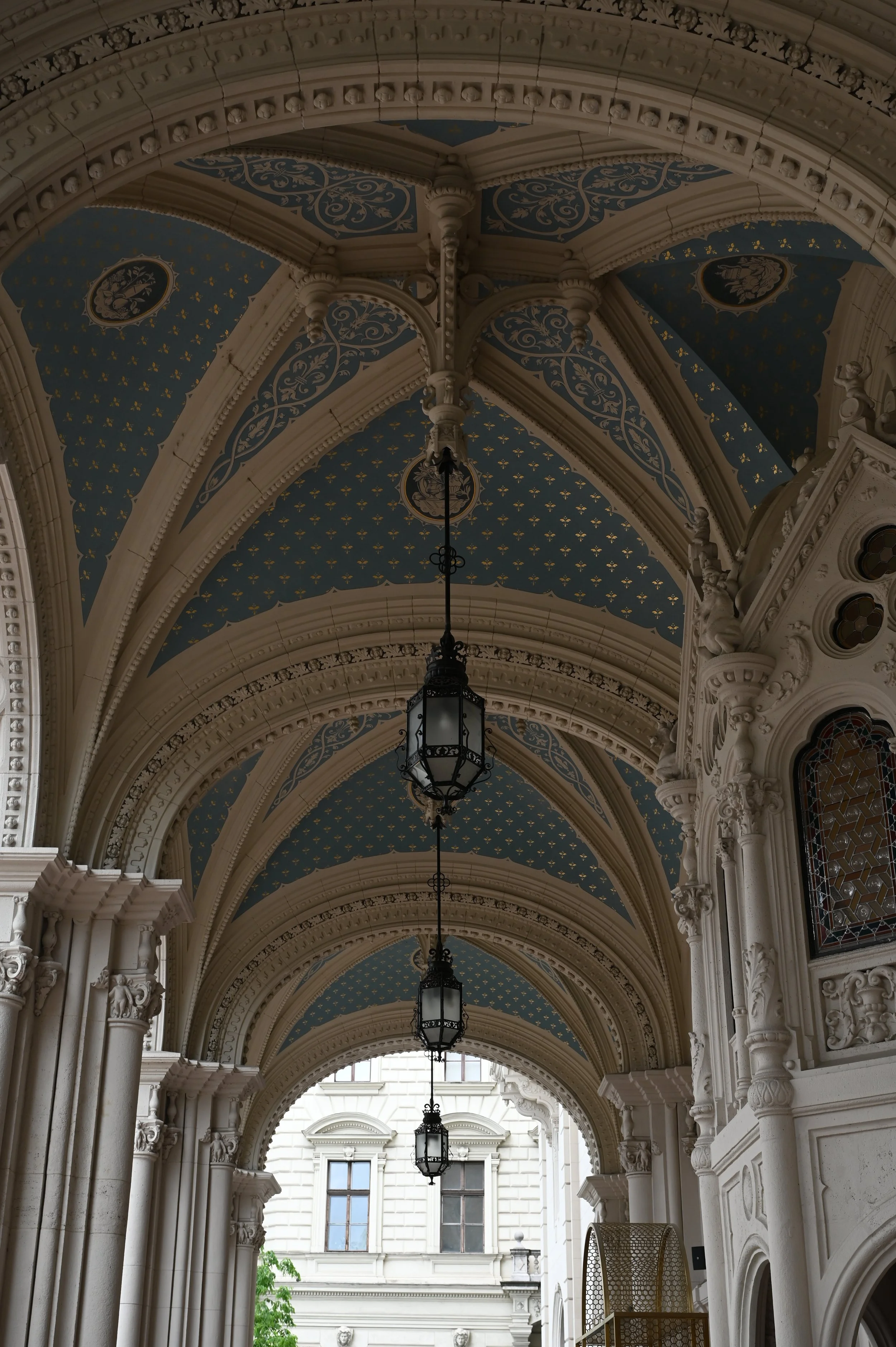 Elegant arched hallway with blue ceiling decorated with gold floral patterns, ornate white columns, and black hanging lanterns.