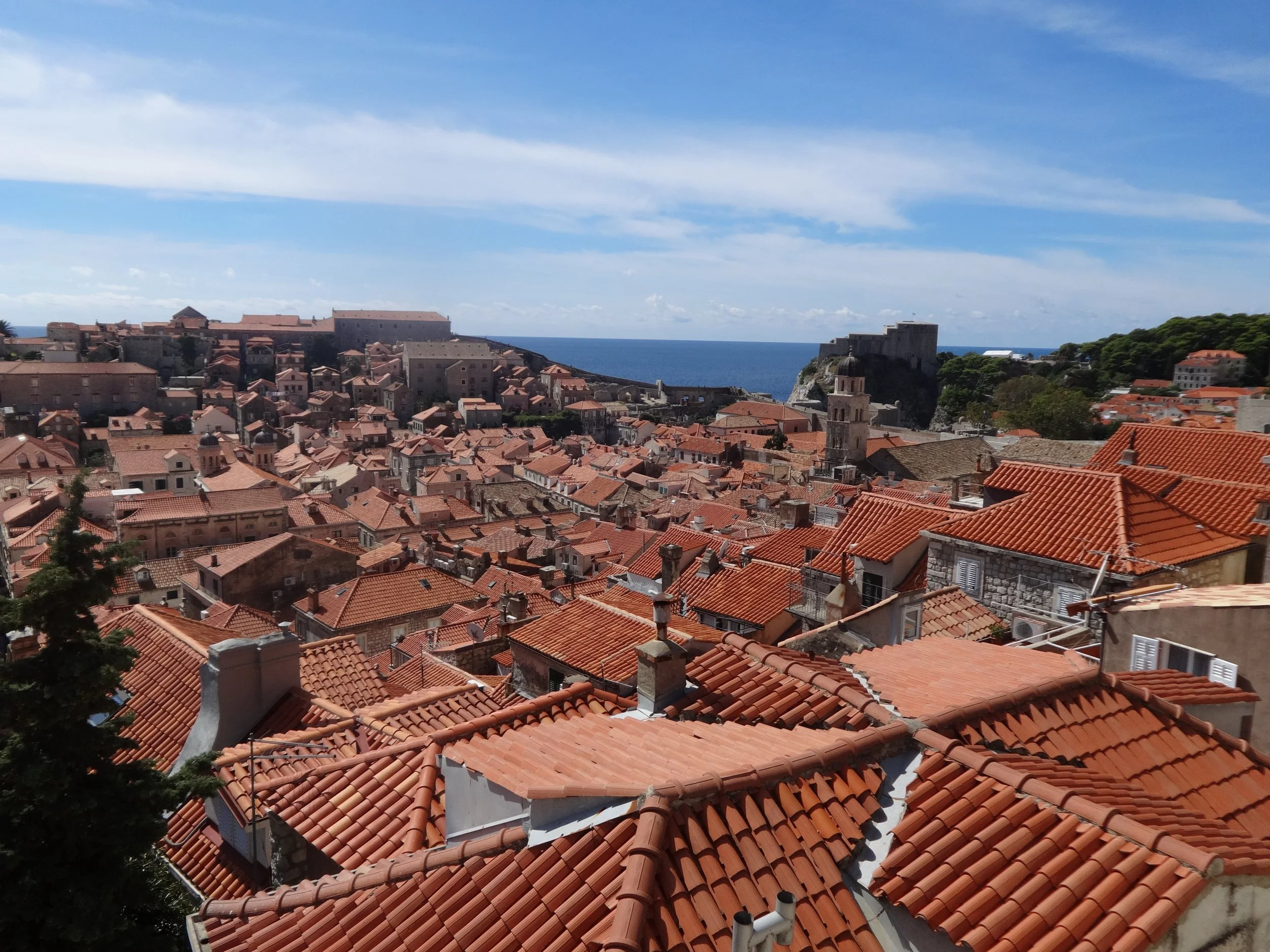A panoramic view of a historic coastal town with terracotta rooftops, a church tower, and the sea in the background under a partly cloudy sky.