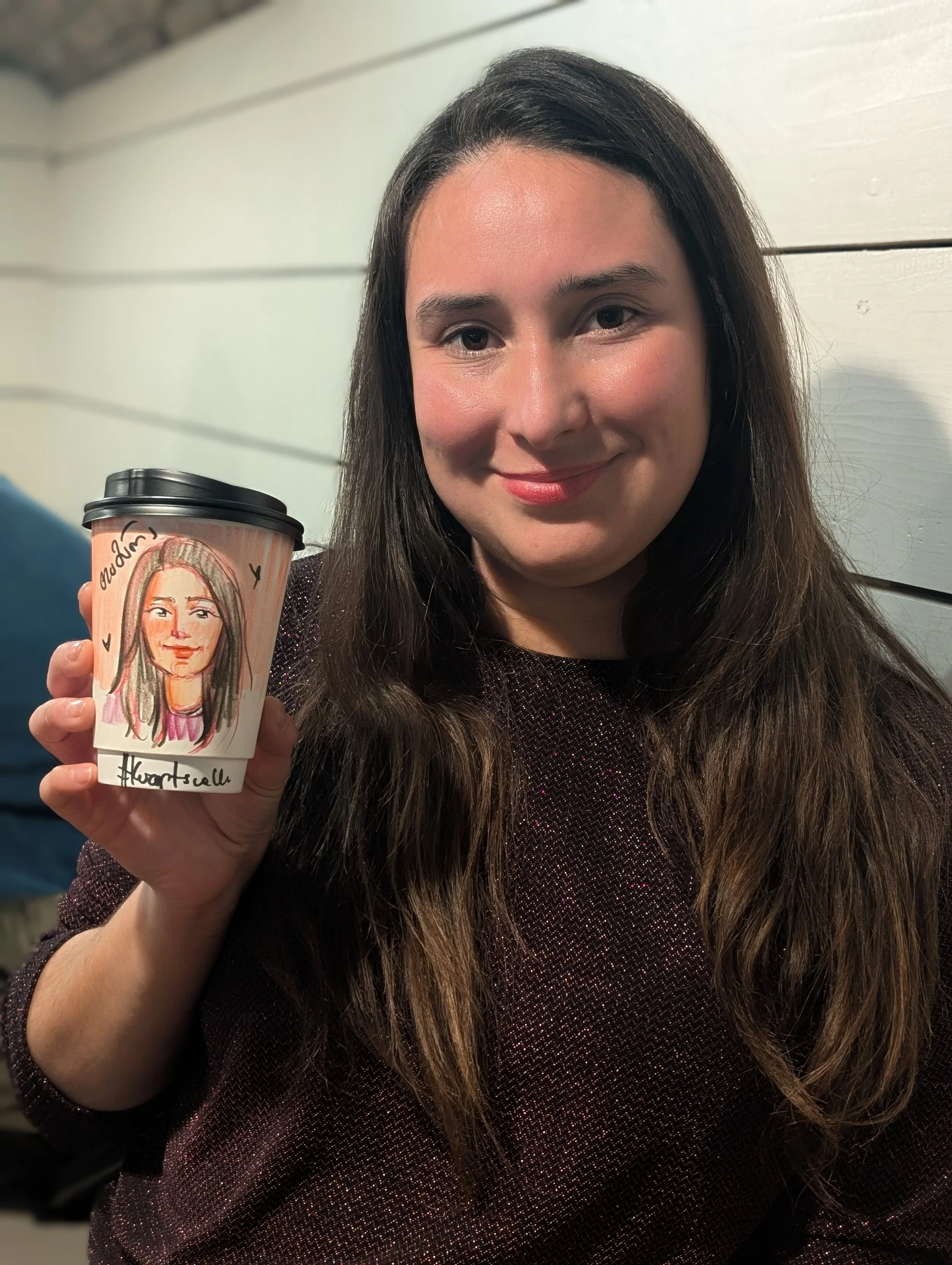 A woman with long brown hair holding a paper coffee cup with a drawn portrait of herself on it, standing in front of a light-colored wooden wall.