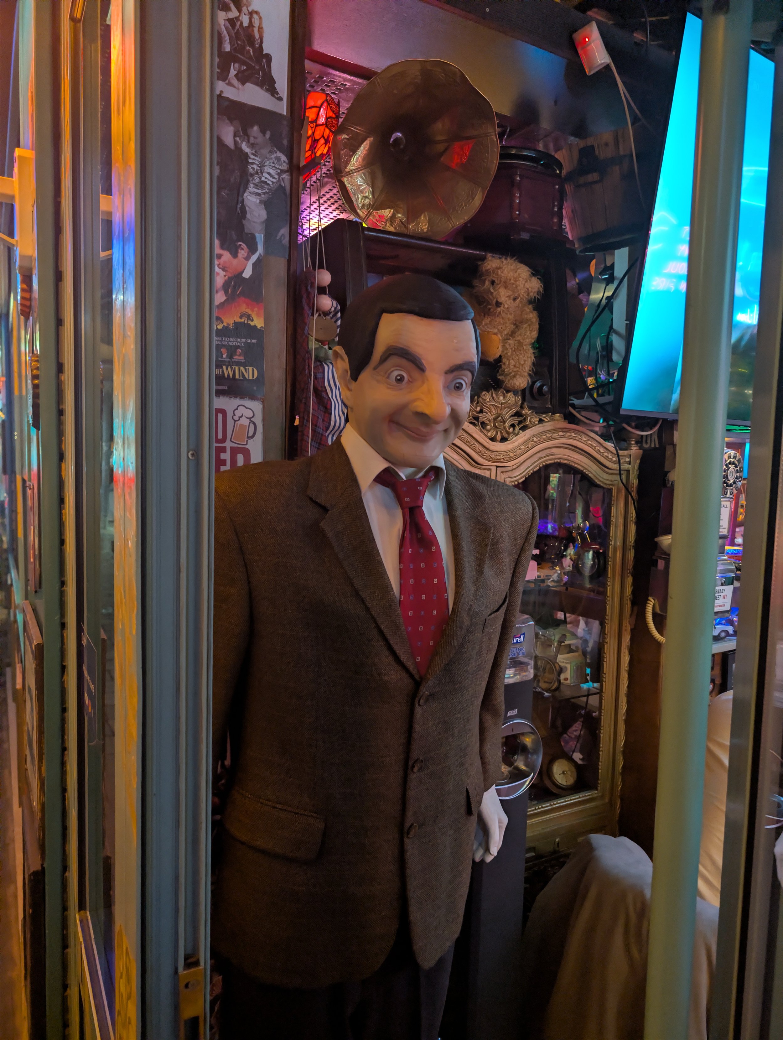 A comic mannequin dressed in a brown suit with a red tie is standing in a room with vintage and nostalgic items, including a photograph, a phonograph, a teddy bear, and a neon sign.
