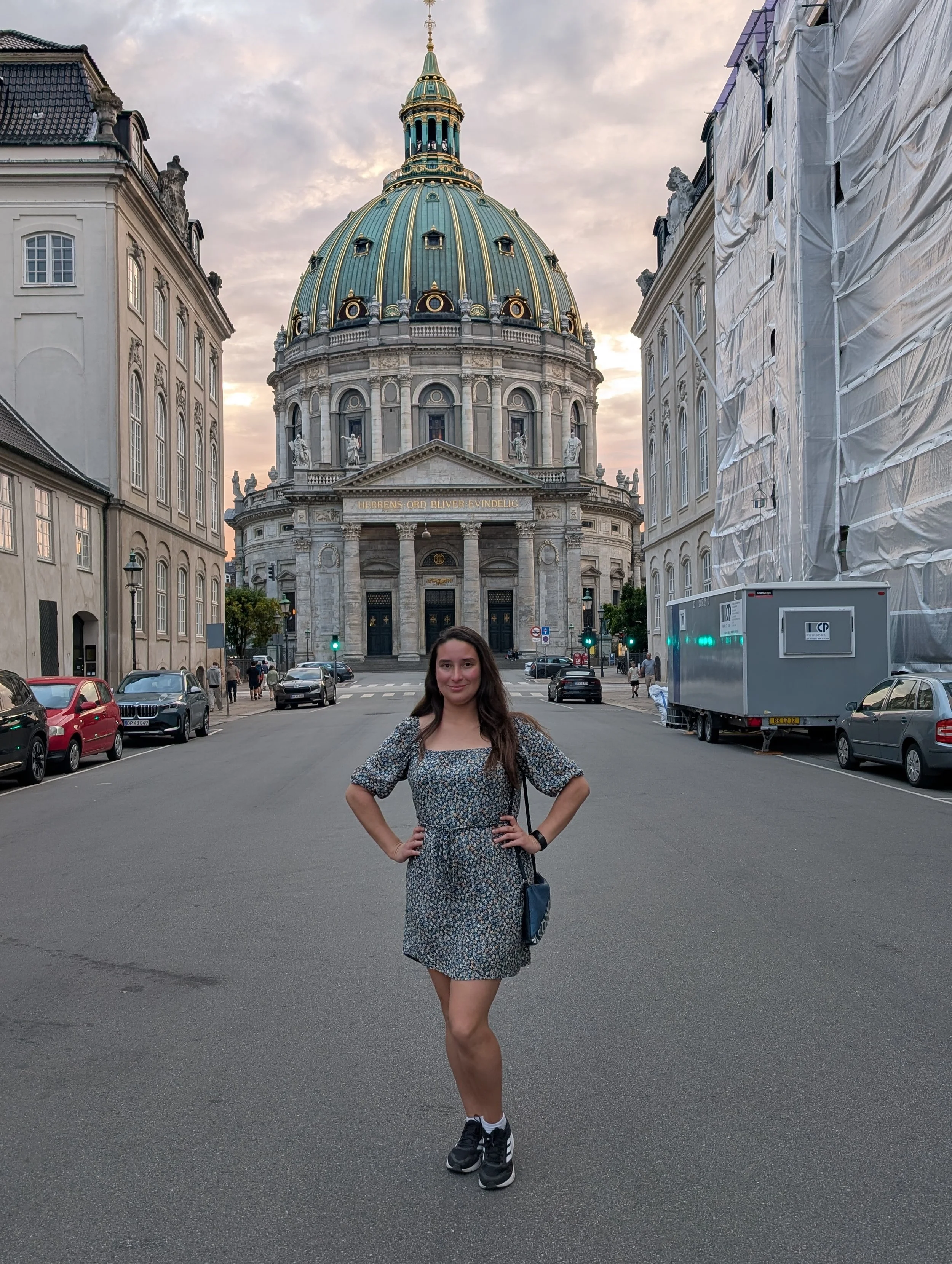 A young woman with long dark hair standing in the middle of a city street with her hands on her hips. She is wearing a patterned dress, sneakers, and carrying a small shoulder bag. Behind her, there is a large historic building with a dome and statue