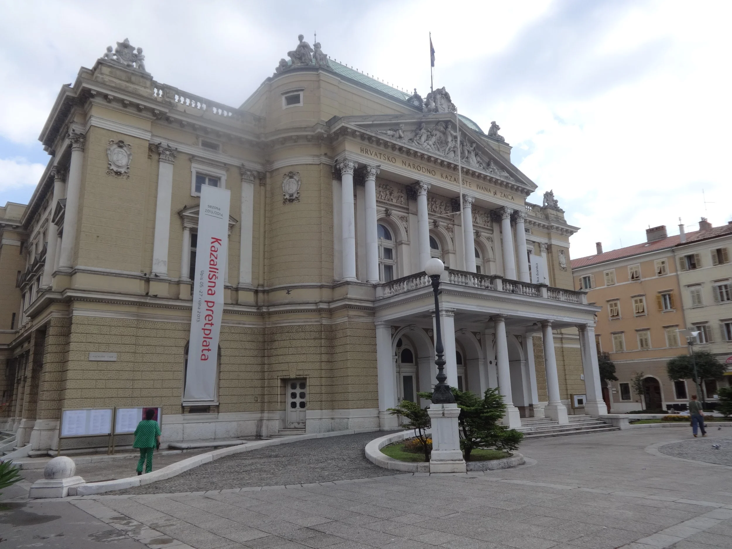 A historic building with ornate architecture, columns, statues, and decorative carvings, with a flag on top, and a few people walking nearby in an urban setting.