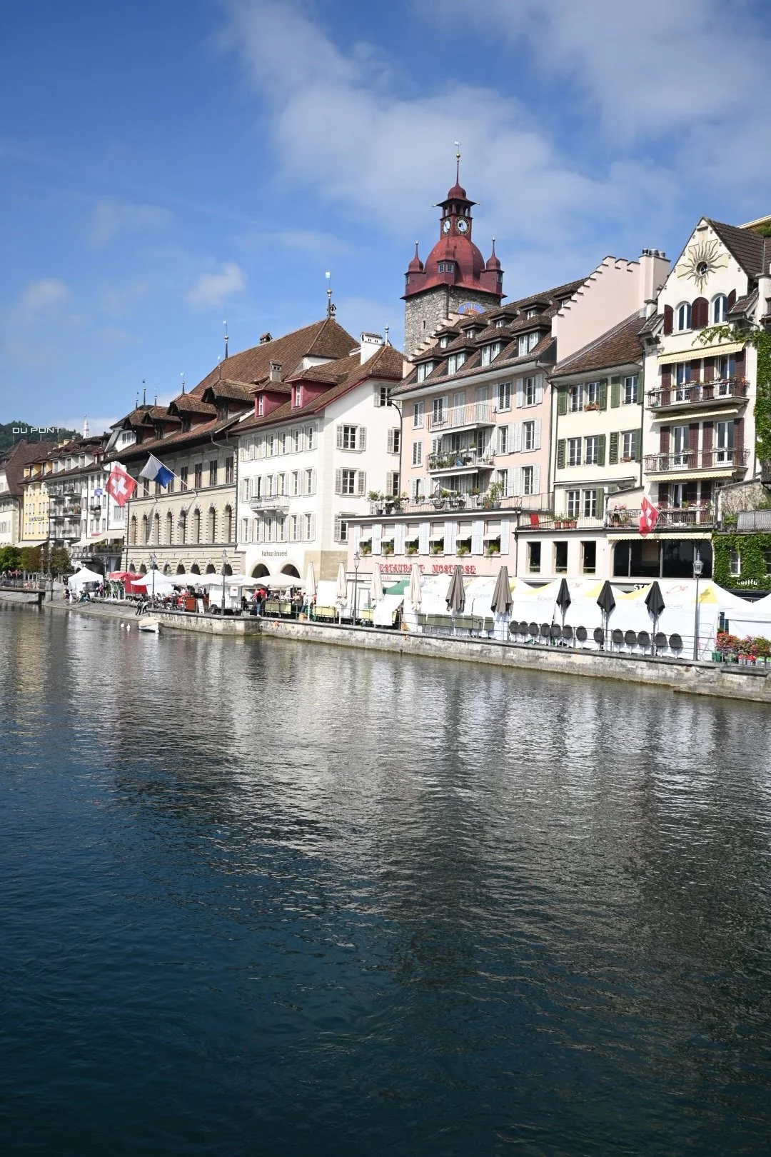 Scenic view of a riverside European town with colorful buildings, outdoor cafes with umbrellas, and a clock tower against a partly cloudy sky.