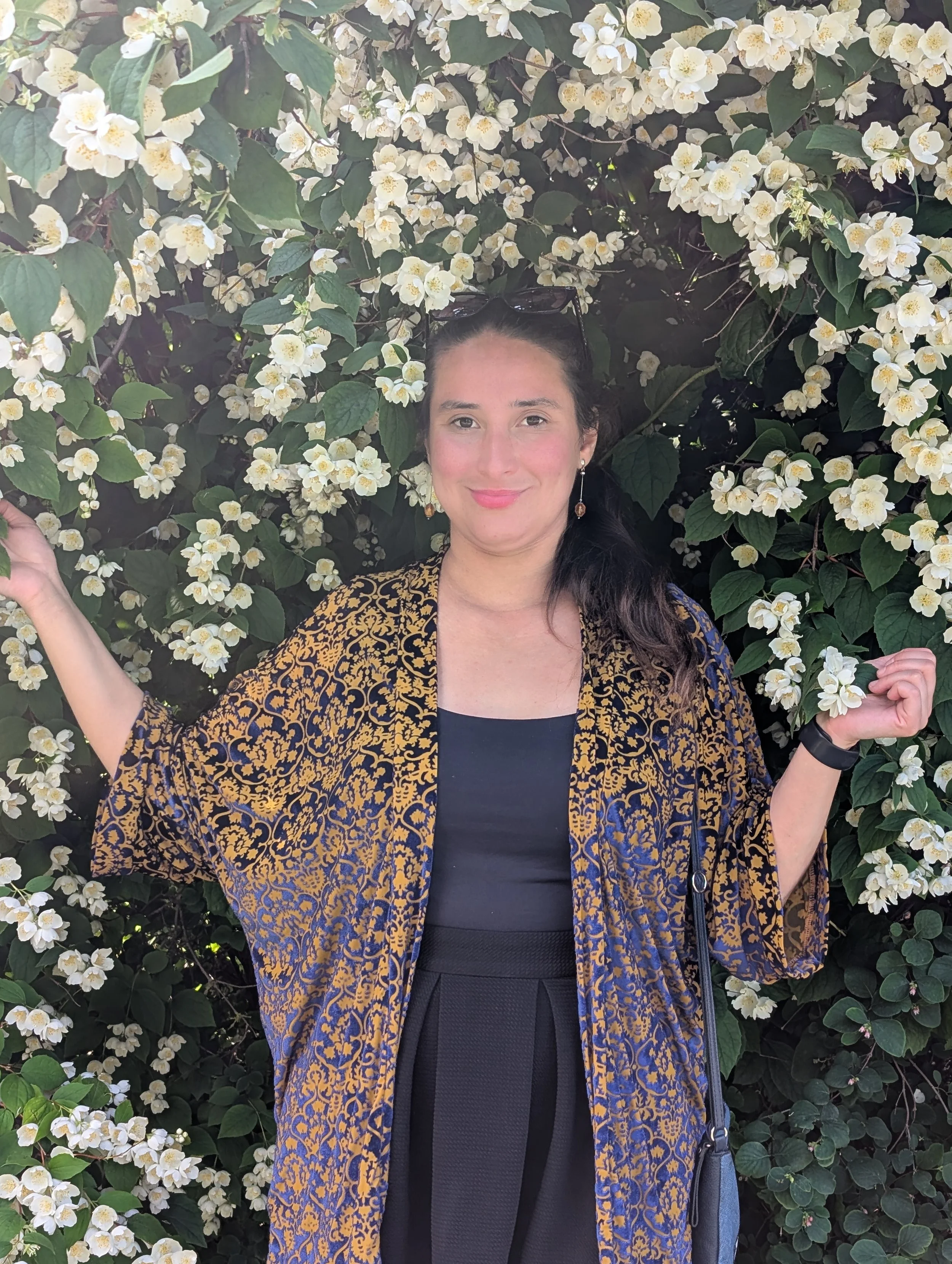 A woman smiling in front of white flowering bushes, wearing a patterned blue and gold jacket, black top, and earrings, holding sunglasses on her head and a black purse.