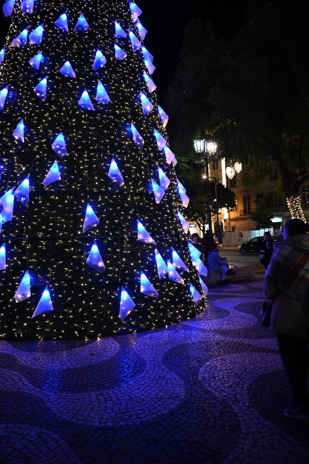 Decorated Christmas tree with blue cone-shaped ornaments and string lights at night in an urban square.