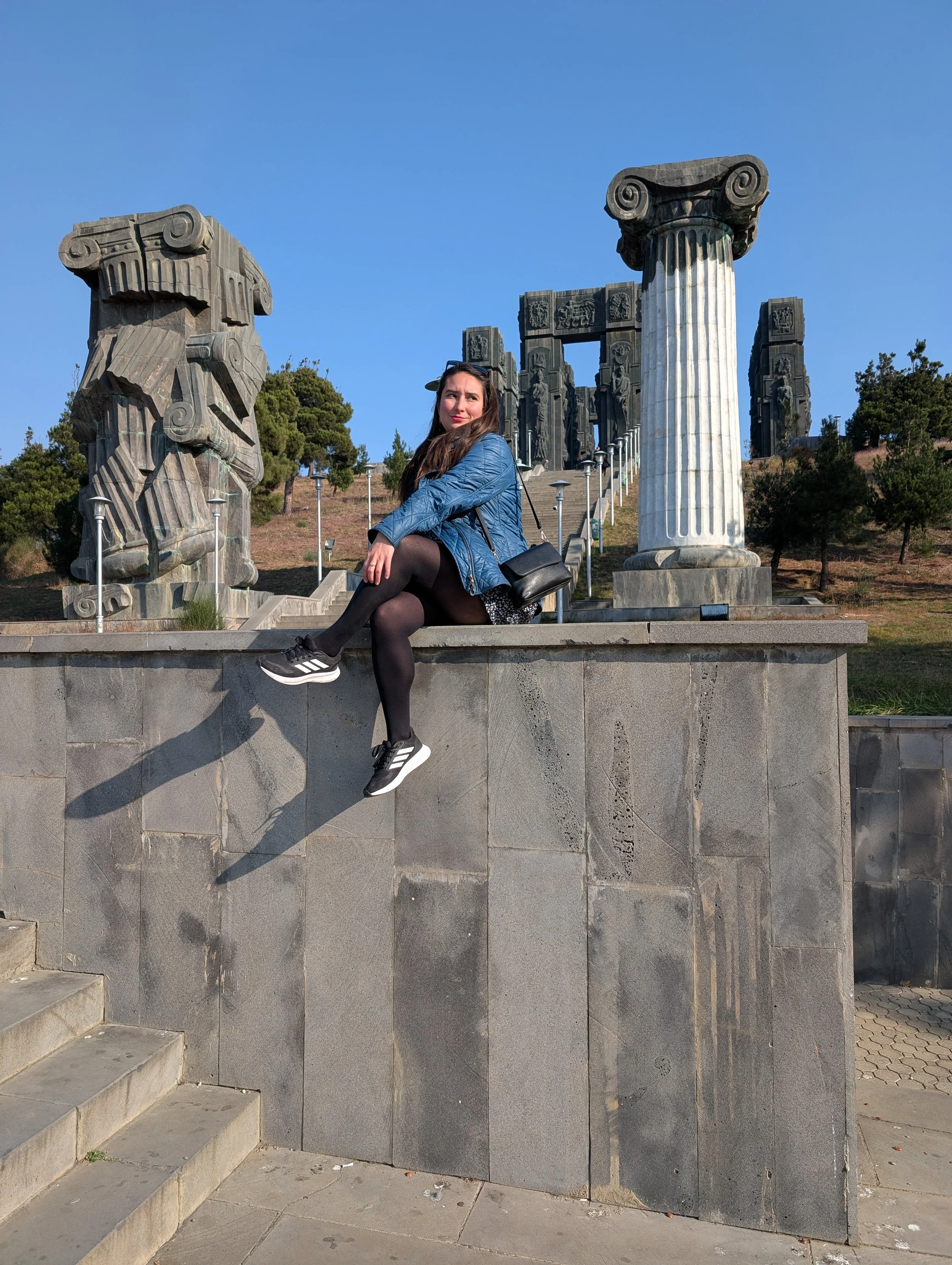 A woman sitting on a stone wall in front of ancient-style stone columns and ruins, with stairs behind her, trees, and a clear blue sky.