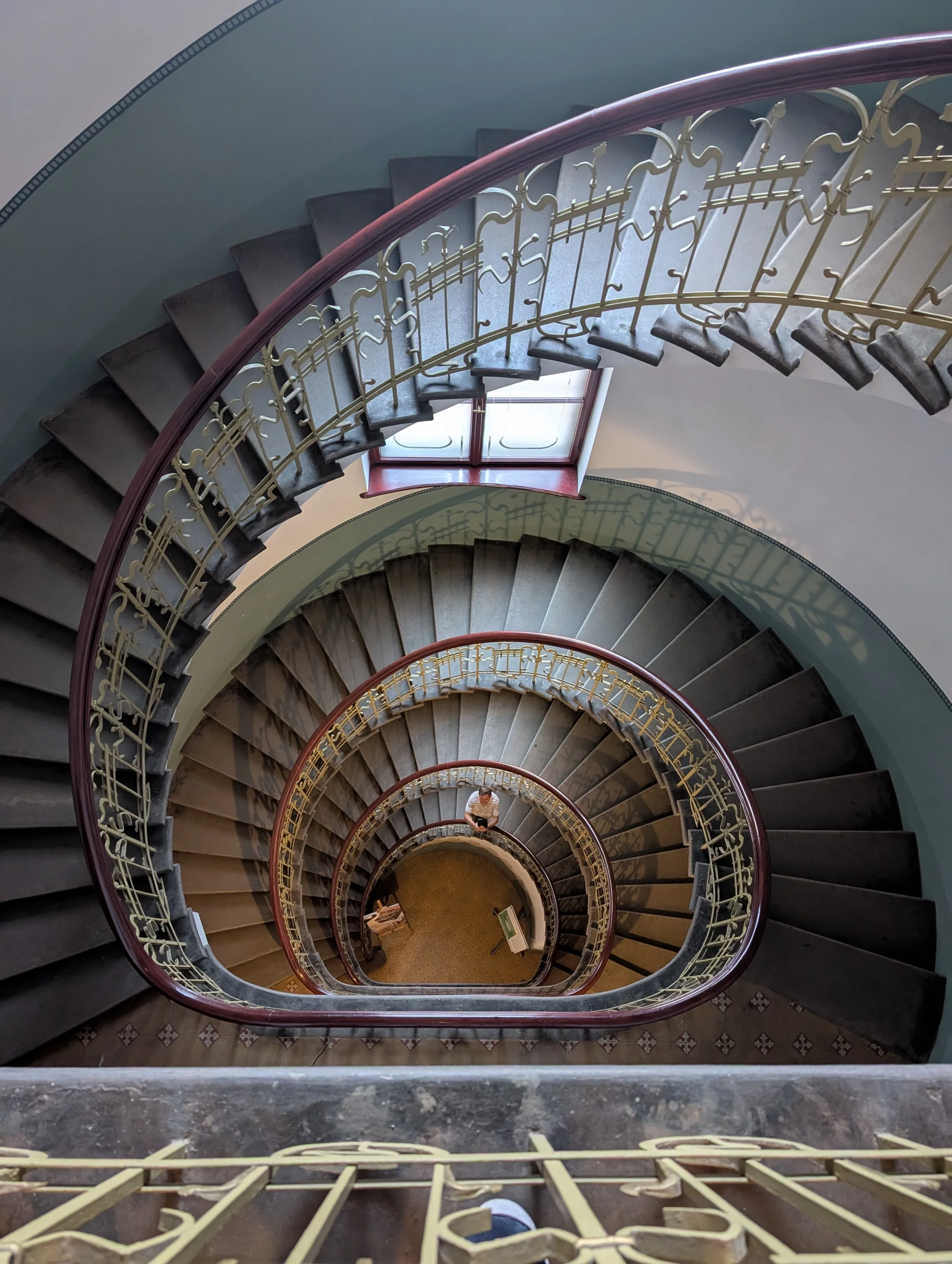 Top view of a winding staircase with metal railing and dark steps, leading downward through multiple floors. A person is sitting at the bottom of the staircase.