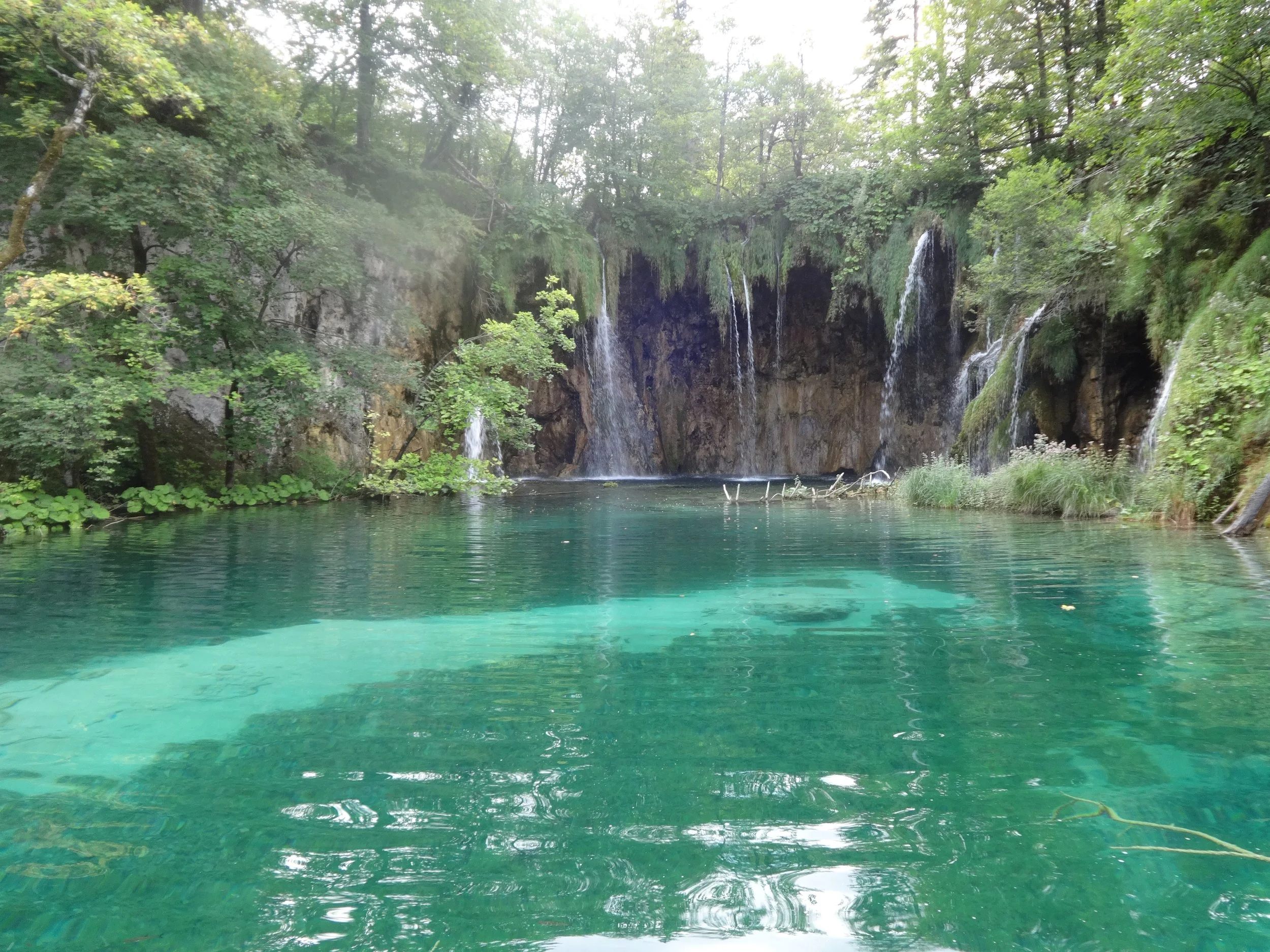 A scenic view of a lush green landscape featuring waterfalls cascading into a clear blue-green body of water, surrounded by dense trees and foliage.
